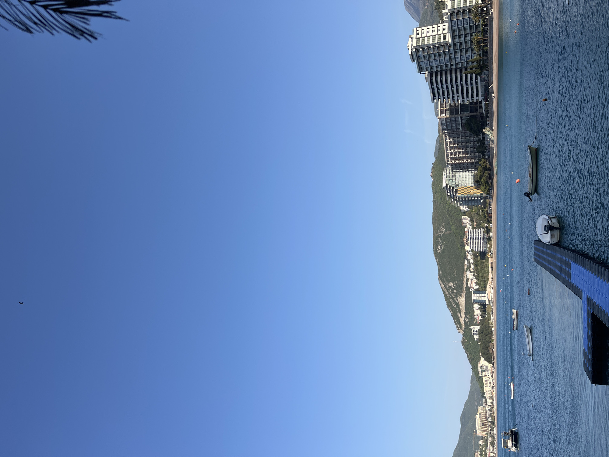 The image presents a bright, sunny coastal scene, likely Budva, Montenegro, viewed from a slightly elevated position over the water. The composition is horizontally dominated by a calm, rippled blue sea on the right, and a clear, cerulean sky on the left.
In the foreground, a dark silhouette of palm fronds is visible at the top left. A prominent feature in the water on the right is a blue and black modular floating dock or platform, extending towards the viewer. Several small boats are moored or floating in the tranquil water, including a light-colored motorboat near the dock and a darker, longer vessel further out. Orange buoys are scattered across the water, indicating mooring areas or swimming zone boundaries.
The shoreline is visible along the left side of the body of water, featuring a narrow strip of beach. Behind the beach, numerous modern, multi-story buildings with balconies and large windows line the coast. These buildings, appearing to be hotels or residential complexes, are nestled at the base of lush green mountains that rise steeply into the background. More urban development, including buildings and roads, can be seen winding up the lower slopes of the mountains.
The sky is a vibrant, cloudless blue, transitioning subtly to a lighter shade near the horizon. A small, dark bird is captured in flight in the upper-left quadrant of the sky.
The scene depicts a peaceful, clear day, suggesting a holiday or resort setting. The strong sunlight indicates it is likely morning or early afternoon. No specific human activity is discernibly detailed, but the boats and buildings imply recreational and residential use of the area. No visible text is present in the image.