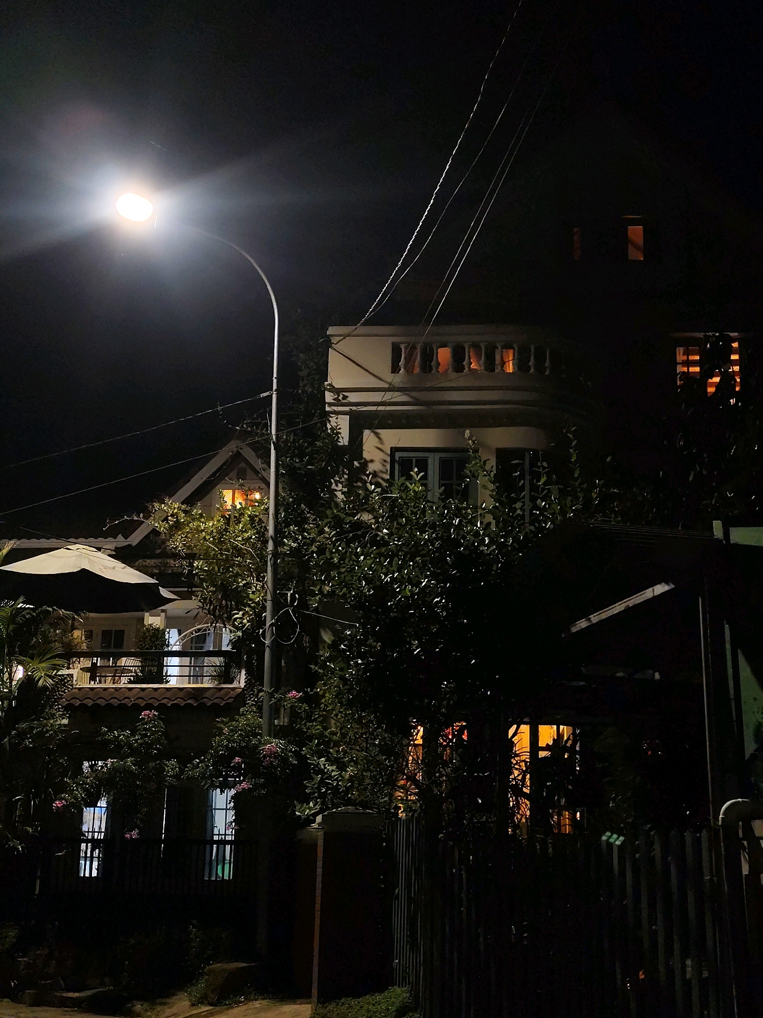 The image depicts a street scene at night in Dalat, Vietnam. A bright street lamp illuminates the foreground, casting a strong glow on its curved pole and the surrounding darkness. In the background, a multi-story building with illuminated windows is visible. The building's architecture suggests a colonial or French-influenced style, common in Dalat. Lush green foliage and trees partially obscure the building, creating a sense of depth and shadow play. A black wooden fence runs across the lower portion of the image, separating the street from the property. The overall atmosphere is tranquil and slightly mysterious, characteristic of a quiet night in a Vietnamese city known for its cool climate and picturesque landscapes. There are no people or specific actions visible in the scene.