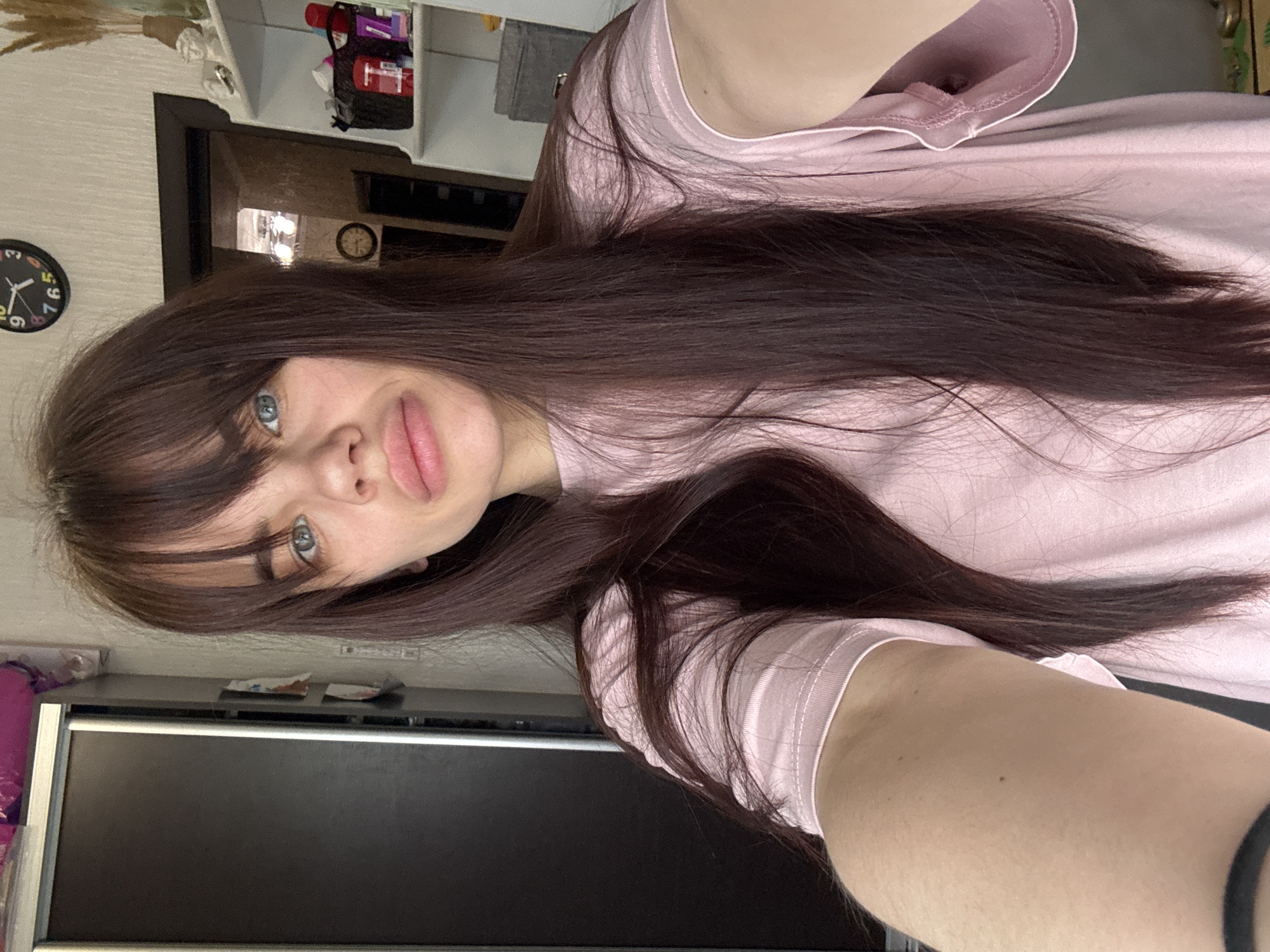 This media file features a young woman taking a selfie in an indoor setting, likely a home, in Rostov-on-Don, Russia. The image is rotated 90 degrees counter-clockwise.
The primary subject is a young woman with long, straight, dark brown hair, styled with bangs across her forehead. She has light blue or grey eyes and full lips, wearing a neutral to slightly pouting expression while looking directly at the camera. She is dressed in a plain, light pink t-shirt. A black elastic hair tie is visible on her right arm.
In the background, on the left, a black round wall clock with white numbers and hands shows the time as approximately 1:55. Above the woman's head, a white wall-mounted shelf unit holds various items, including a black bag with a red "T" logo, possibly containing toiletries or makeup, and some decorative objects. Behind her, a dark doorway or opening leads to another room, reflecting what appears to be an overhead light fixture. A second, smaller clock is faintly visible on the wall through the doorway. In the lower right portion of the image, a large, dark electronic screen, likely a computer monitor or television, rests on a dark surface. Several small pieces of paper or wrappers are visible on the surface near the screen. The walls are a light, textured color.
The scene conveys a casual and personal atmosphere, typical of a selfie taken at home. The time on the prominent clock suggests it could be early morning or afternoon, but the indoor lighting suggests no specific time of day is strongly indicated beyond general daylight hours.