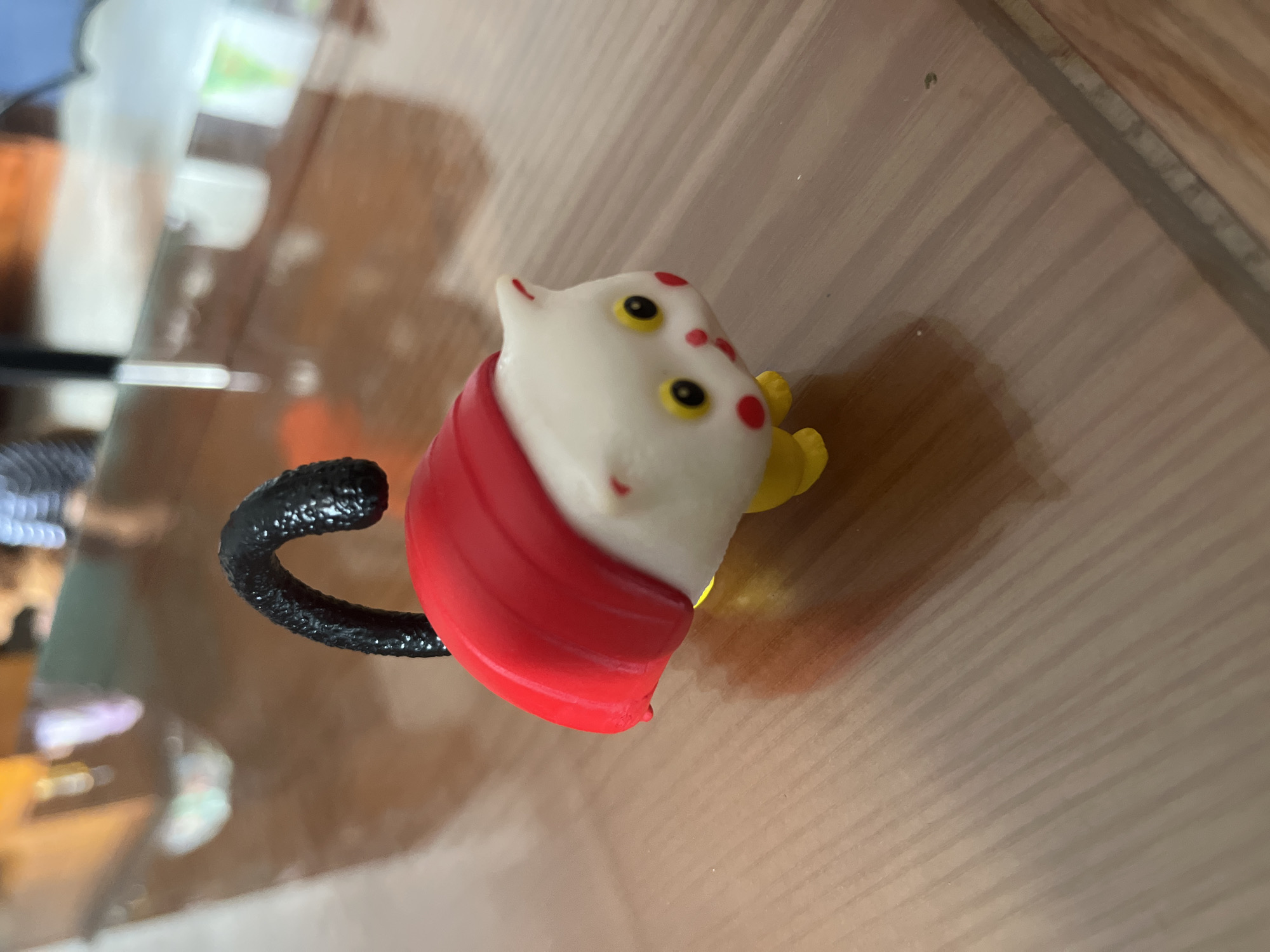 A toy cat figure sits on a wooden surface. The cat is primarily white with yellow eyes and red markings. It wears a red cape and has a black tail. The background is a wooden surface, likely a table or counter. The location and specific details about Bac Nha Trang Ward, Vietnam, are not discernible from the image.