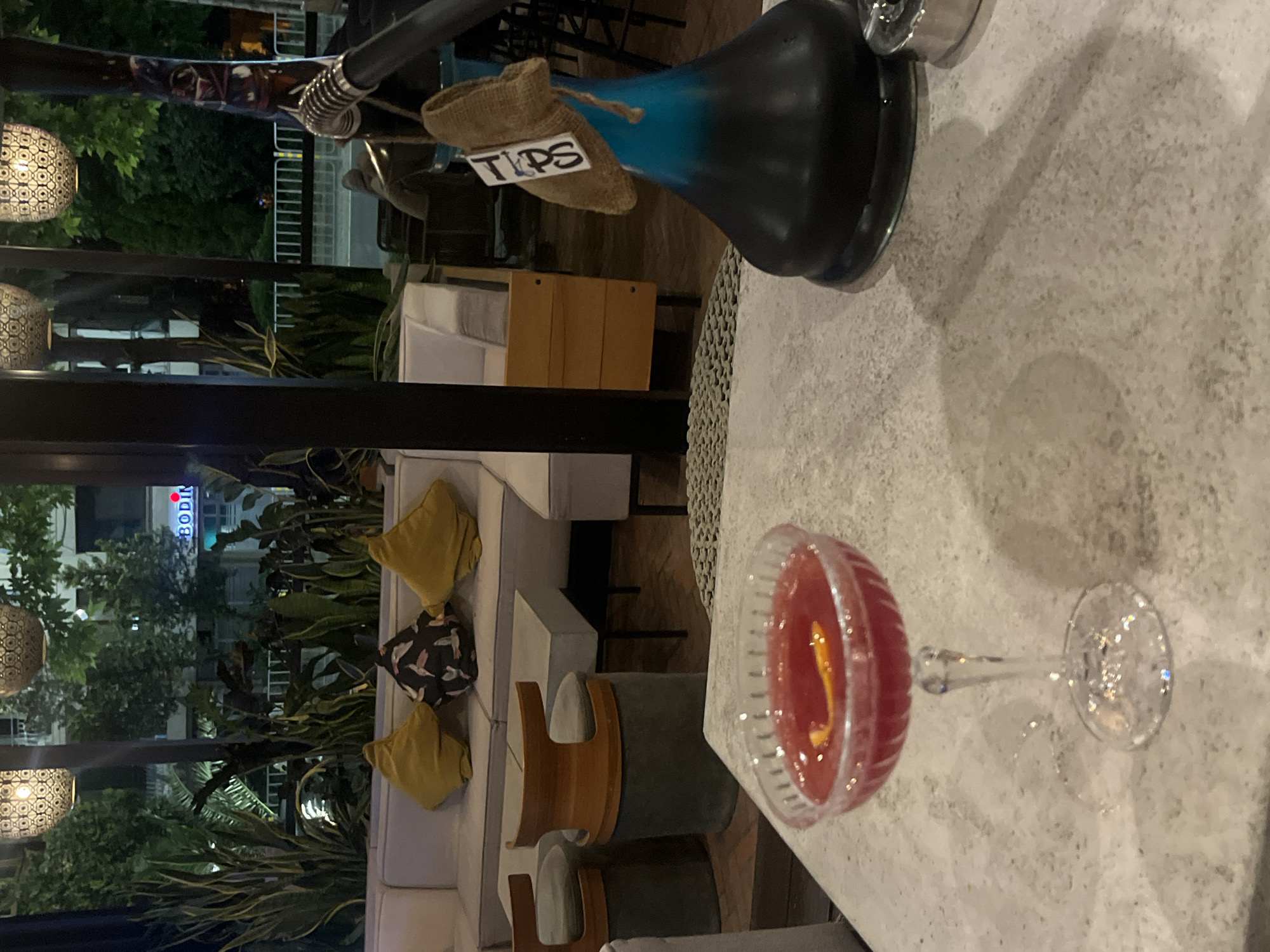 The image depicts an outdoor seating area, possibly at a bar or restaurant, at night. In the foreground, a cocktail is served in a coupe glass on a textured surface. To the right, a shisha pipe with a blue glass base is visible, with a burlap pouch labeled "TIPS" hanging from it. In the background, a white sectional sofa with yellow and patterned cushions is arranged around low tables. Beyond the seating area, there is lush greenery, including palm-like plants, and streetlights illuminate the scene. A street is visible in the distance with some buildings. The overall ambiance suggests a relaxed, evening setting. The text on the shisha pipe, "TIPS," and the presence of an outdoor seating area are consistent with establishments found in Burgas, Bulgaria, particularly in areas with nightlife and entertainment.