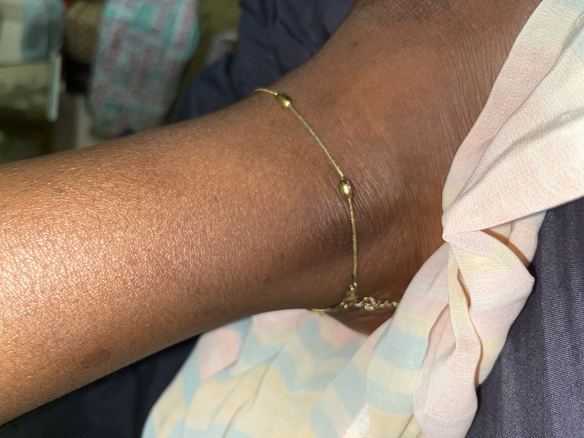 A close-up shot focuses on the ankle and lower leg of a person with dark skin. A delicate gold chain anklet adorns the ankle, featuring small, elongated bead-like charms. The anklet is partially obscured by a pastel-colored fabric with a striped pattern in shades of pink, yellow, and blue, which drapes across the lower leg and foot. The background is out of focus, showing hints of dark fabric and what appears to be a cluttered surface with patterned materials. The lighting is dim, highlighting the texture of the skin and the shine of the gold anklet. The image appears to be taken indoors. The specific location in Abuja, Nigeria, cannot be determined from the visual information alone.