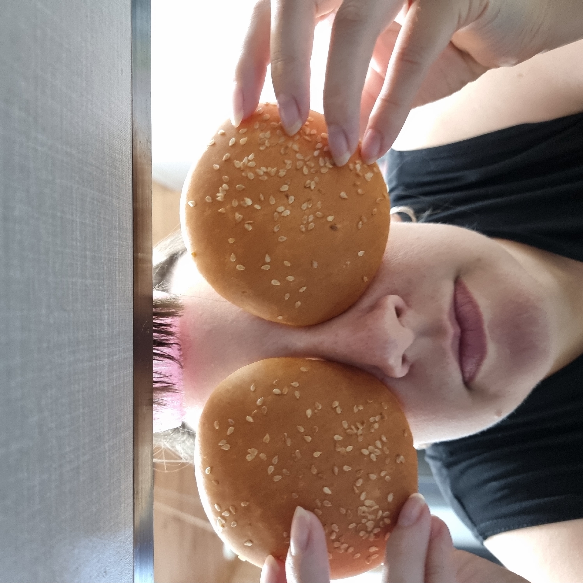 The image shows a person's face from the nose up, with two sesame seed hamburger buns covering their eyes. The person is wearing a black top. The background is blurred, but appears to be indoors with a light-colored surface above. The image does not contain any information about the location in Dunnae-myeon, South Korea.