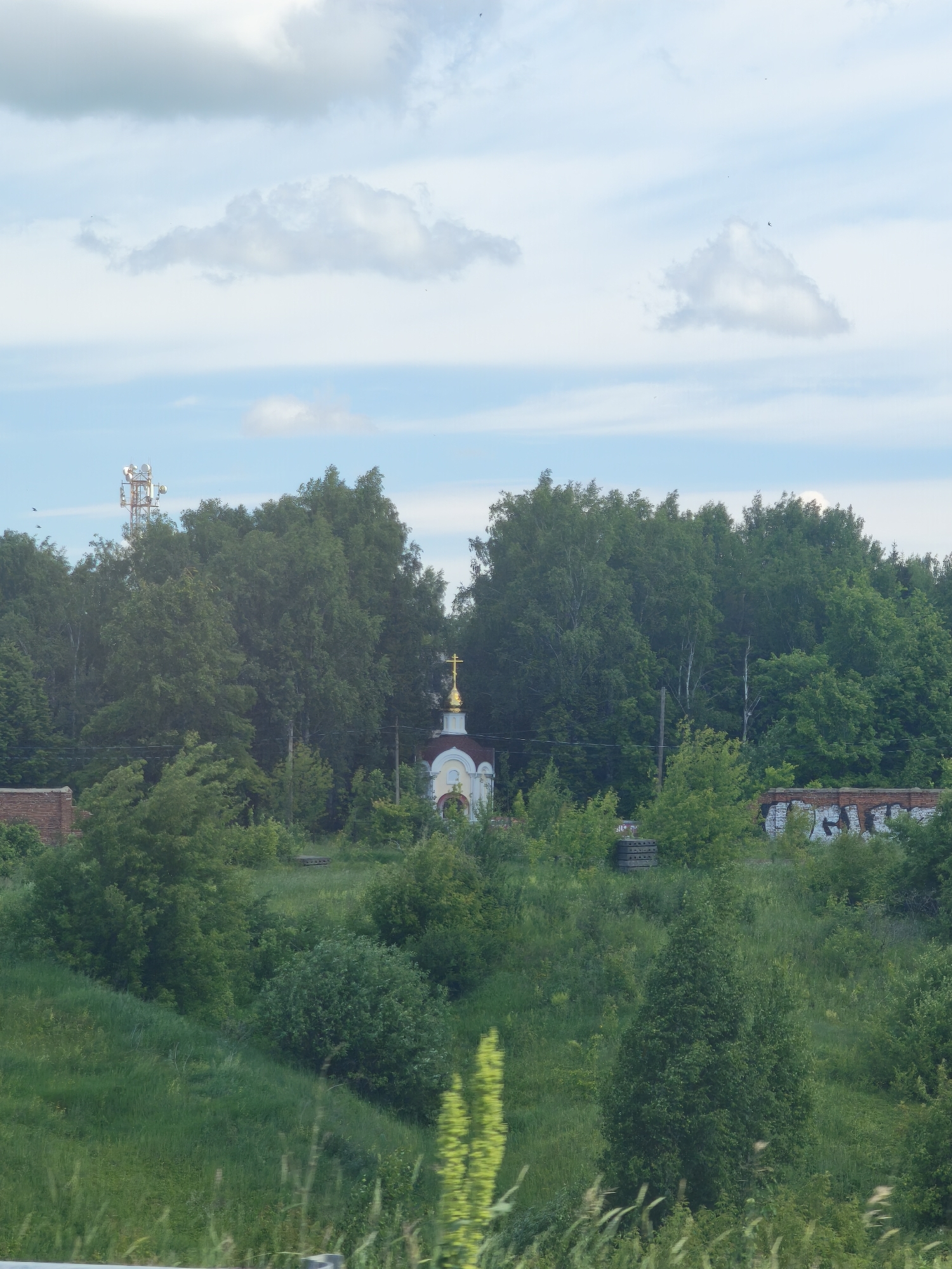 In Kanash, Russia, a small, white and gold-domed chapel stands amidst dense green trees. A red brick wall, partially covered in graffiti, is visible to the right of the chapel. To the left of the chapel, a communication tower rises above the treeline. The sky above is a pale blue, dotted with soft white clouds. The foreground is filled with lush green grass and vegetation.