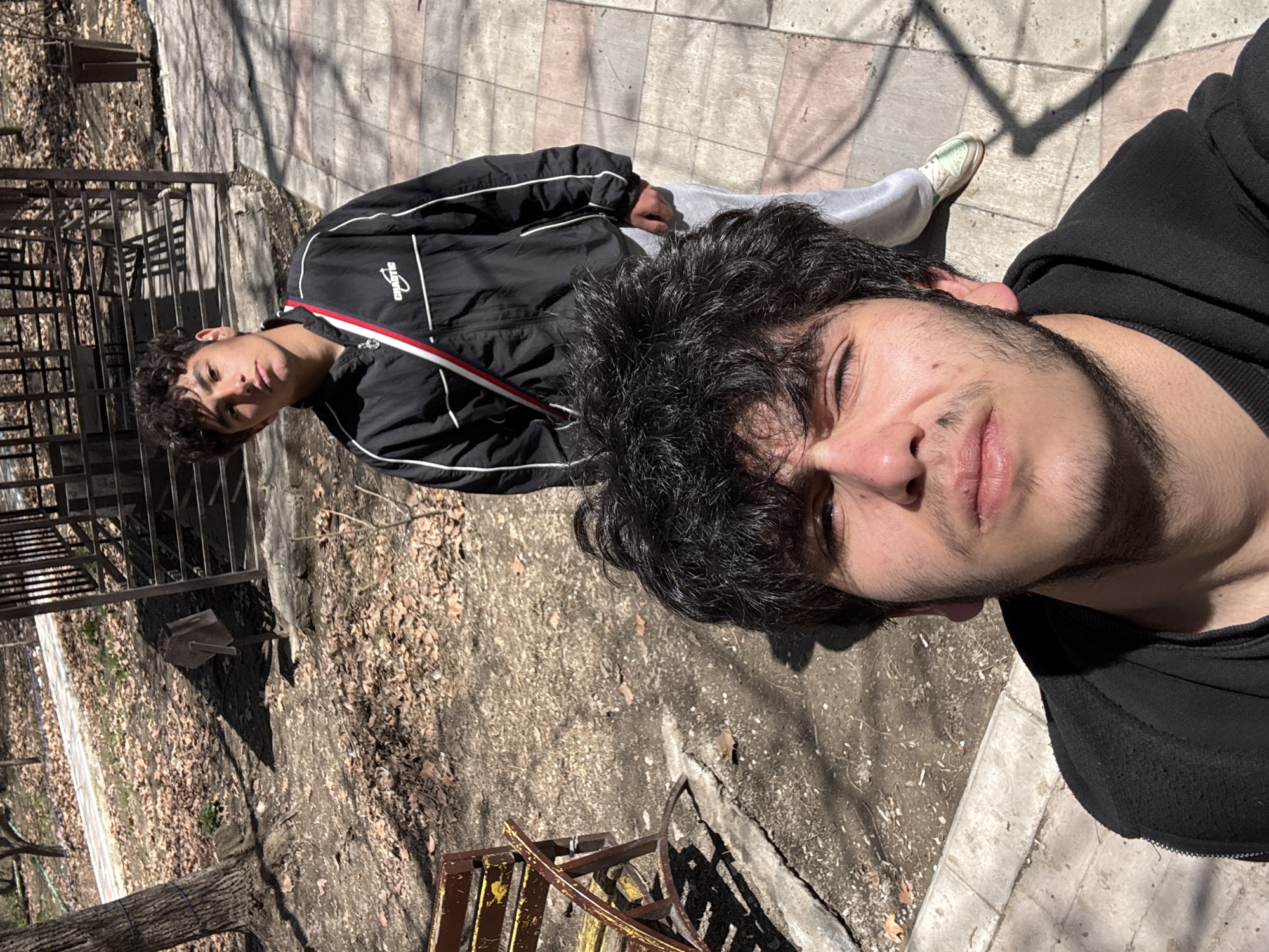 The image shows two young men standing outdoors on a partly sunny day. They are positioned on what appears to be a sidewalk made of light-colored paving stones. Behind them is an area of bare ground covered in fallen leaves, and a metal railing. One of the men, who is closer to the camera, is wearing a black hooded jacket. The other man is wearing a black jacket with white and red detailing. The location appears to be a city park, likely in Yerevan, Armenia, based on the architectural style and general urban environment.