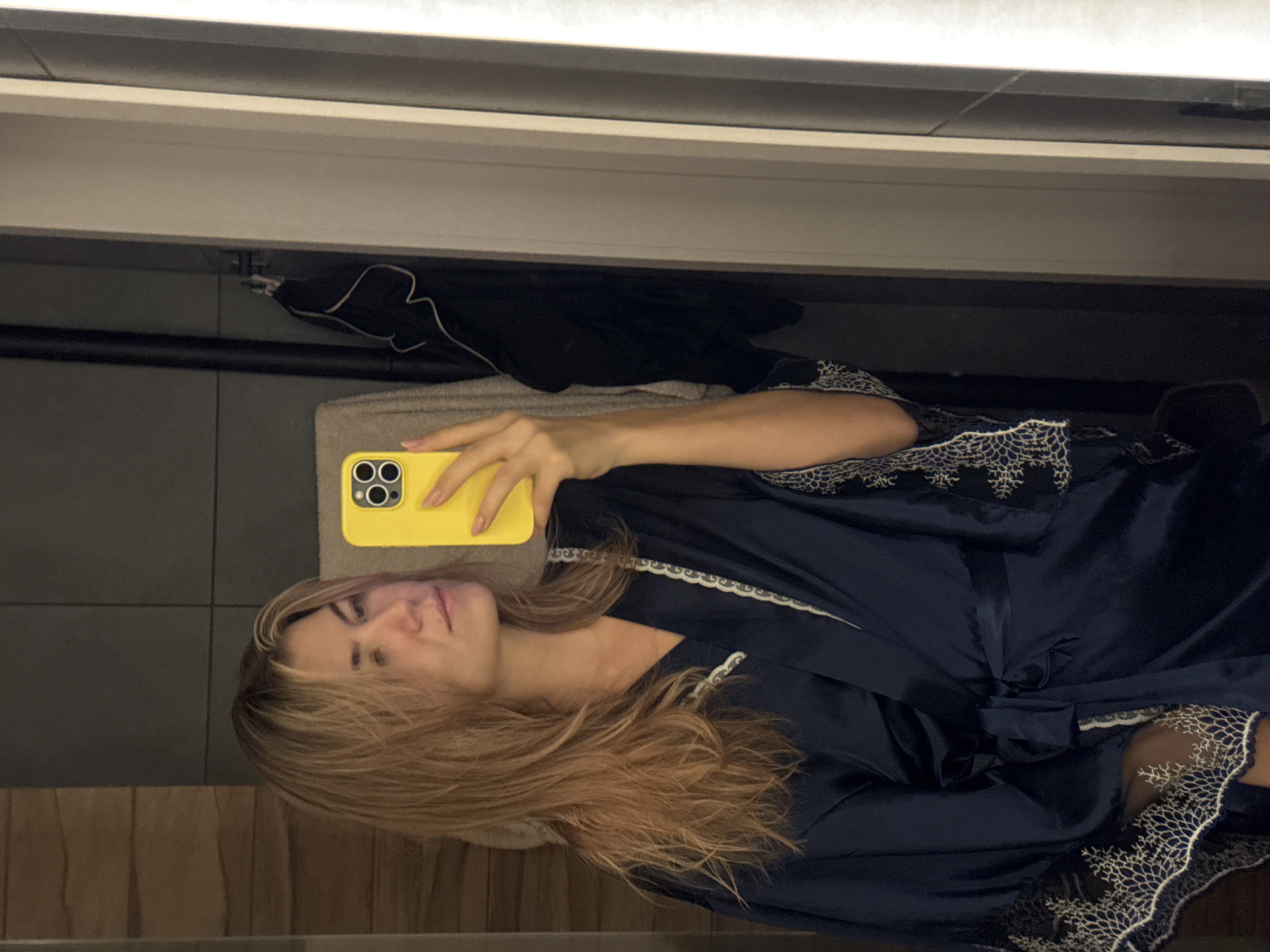 A mirror selfie of a woman in a blue robe with white lace detail, holding a yellow iPhone. The location appears to be a bathroom. There is black clothing hanging on a towel bar in the background. The city and state are unknown.