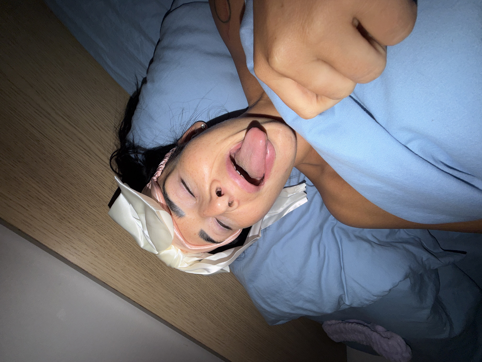 A woman is lying in bed under a light blue duvet. She has a satin sleep mask on her forehead, a pink headband, and her eyes are closed. Her mouth is open, and her tongue is sticking out. Her right hand is visible near her head, partially covered by the duvet. The scene appears to be indoors, likely a bedroom. A portion of a wall or furniture with a light wood finish is visible in the background on the left. A pink slipper is partially visible on the bottom of the image. No information indicates a location in Saint Petersburg, Russia.