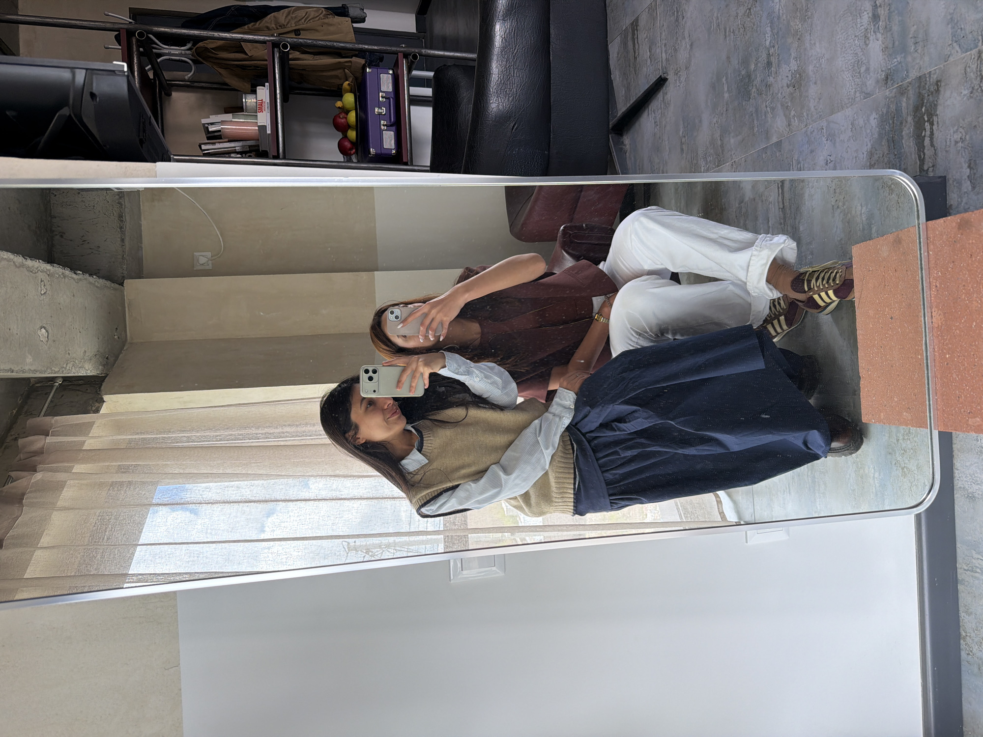 An indoor mirror selfie features two young women in Yerevan, Armenia, captured during the daytime. The image shows the women reflected in a large mirror.
The woman on the left in the reflection has long dark hair and wears a light blue and white striped shirt layered under a beige V-neck sweater vest, paired with a dark blue long skirt. She holds a light-colored smartphone (likely an iPhone) with her right hand, actively taking the photo, with a slight smile on her face.
The second woman, to her right, also has long dark hair. She wears a dark reddish-brown top or jacket, white wide-leg trousers, and brown and white striped sneakers (resembling Adidas Gazelle). She is also holding a light-colored smartphone, looking at its screen, with her face partially obscured.
The reflection reveals the interior of the room. Behind the women, there's a light-colored wall and a window covered by translucent beige blinds, through which blue sky and clouds are visible, indicating daylight. To the right, a concrete wall with a power outlet is seen. Above the women, a black metal shelving unit holds various items including books (one with "SMALL" visible on its spine), a brown fabric item, a black object, and a small purple suitcase. A bowl of red and green apples is also on the shelf. Part of a dark brown leather armchair is visible, and the floor appears to be light grey concrete or tile, with a terracotta-colored trim at the bottom right of the mirror.