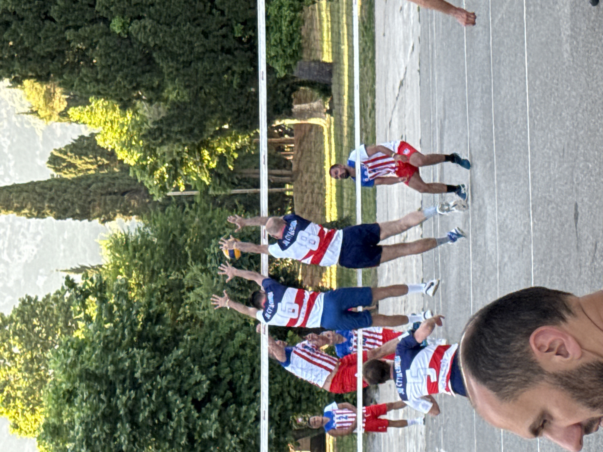 The image depicts a volleyball match in progress on an outdoor court, likely located in Budva, Montenegro, given the context. The court appears to be asphalt, marked with white lines. A white net divides the court. Seven young men, all wearing similar red, white, and blue striped jerseys and shorts, are visible. Three of them are actively engaged in a play at the net, with two jumping to block a volleyball, which is in the air between them. Another player is positioned to receive the ball. Two other players are on the opposite side of the court, one in a ready stance and another standing near the net. A seventh player is visible in the background on the left, standing behind the court. The players' attire suggests they are part of a team or a tournament. Lush green trees form a dense backdrop, with the subtle hint of distant hills or mountains under a bright sky, suggesting daytime and pleasant weather. The overall atmosphere appears to be casual and sporty.