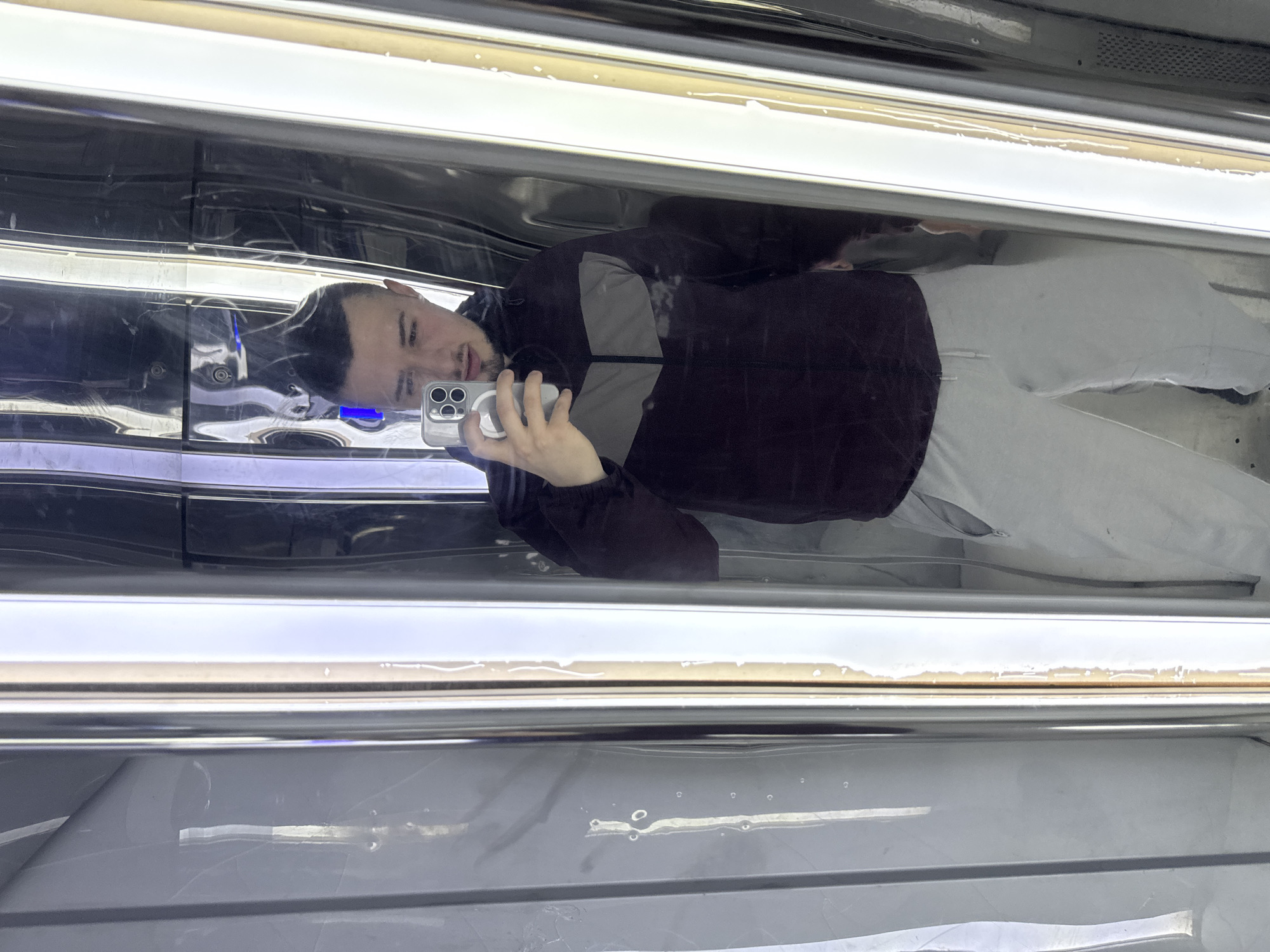 The image appears to be a selfie taken by a man in an elevator or similar enclosed space with mirrored walls. The man is wearing a dark-colored jacket with grey accents and light-colored pants. He is holding a smartphone, presumably to take the picture. The environment consists of reflective panels that create a distorted image of the man and the surroundings. There is no concrete evidence to connect the location to the city of Sofia, Bulgaria, so it is not possible to describe it further.