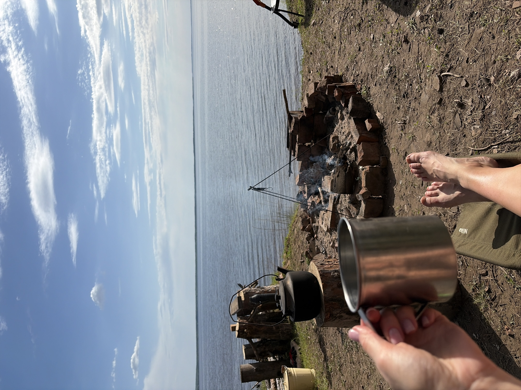 The image depicts a lakeside camping scene. A person is holding a metal mug in their hand, with a view of a lake and cloudy sky in the background. A campfire is burning with smoke, and a black kettle is placed nearby. The ground is covered with dry leaves and grass. A person's feet are visible in the upper right corner. The location is Zabegaevo, Russia.