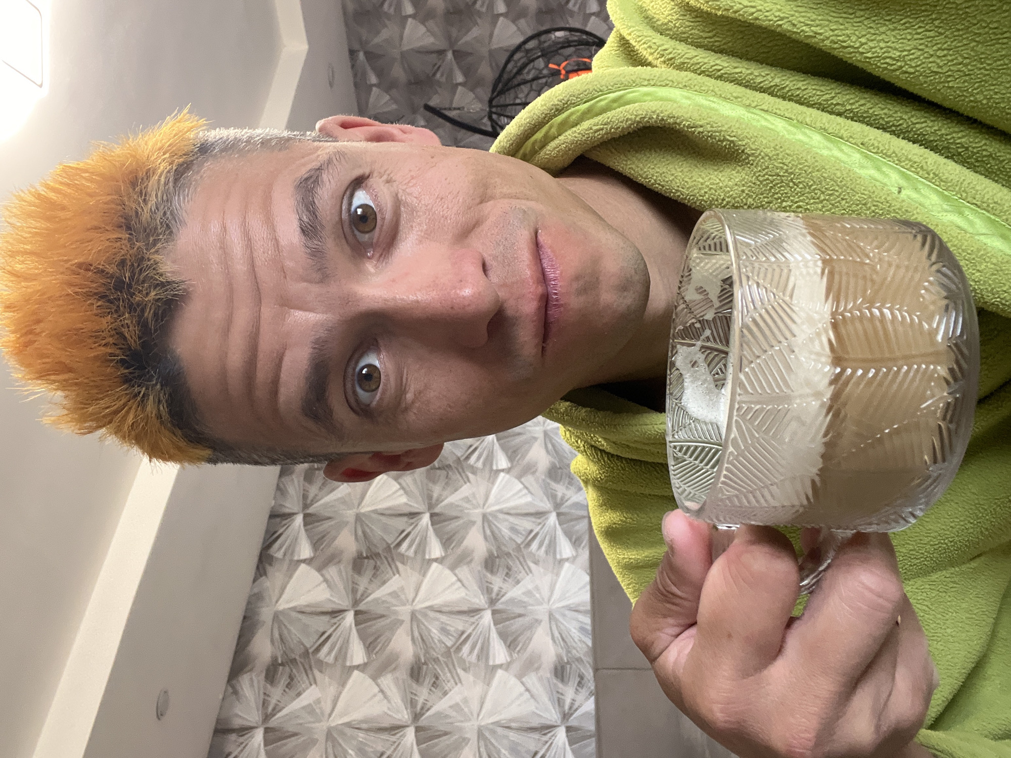 A man with dyed orange hair stands in front of a textured grey and white wall. He is wearing a bright green robe and holding a glass mug filled with a light brown beverage topped with foam. The man's eyes are wide, and he is looking directly at the camera. The mug has a textured pattern of leaf-like shapes. In the background, a black hanging chair with an orange accent is visible. The scene appears to be indoors, likely in a domestic setting, potentially in the city of Chaikovsky, Russia, given the specific request.