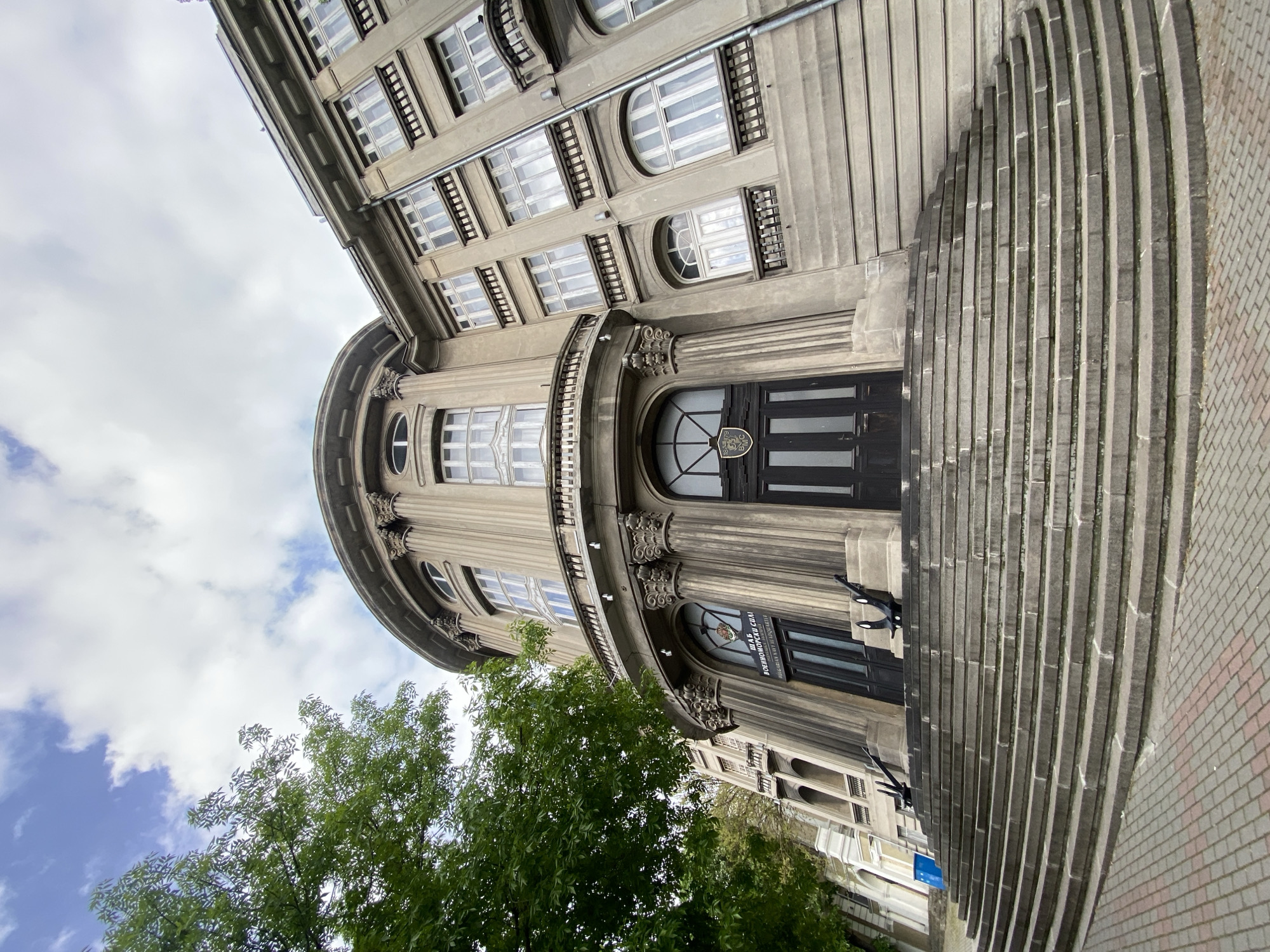 The image shows the Naval Academy "N. Y. Vaptsarov" in Varna, Bulgaria. The perspective is a low-angle shot, looking up at the building's facade and part of the sky. The building has a rounded architectural design, with a classical style evident in the columns and symmetrical window arrangements. A prominent set of steps curves up to the entrance. A tree is visible near the bottom left corner. The sky is partly cloudy. There are a few people near the entrance, including one appearing to be in uniform. The location is an urban environment. The sign on the building reads, in Bulgarian, "Мала военна сала Военноморски сили на Република България" which translates to "Small Military Hall of the Naval Forces of the Republic of Bulgaria."