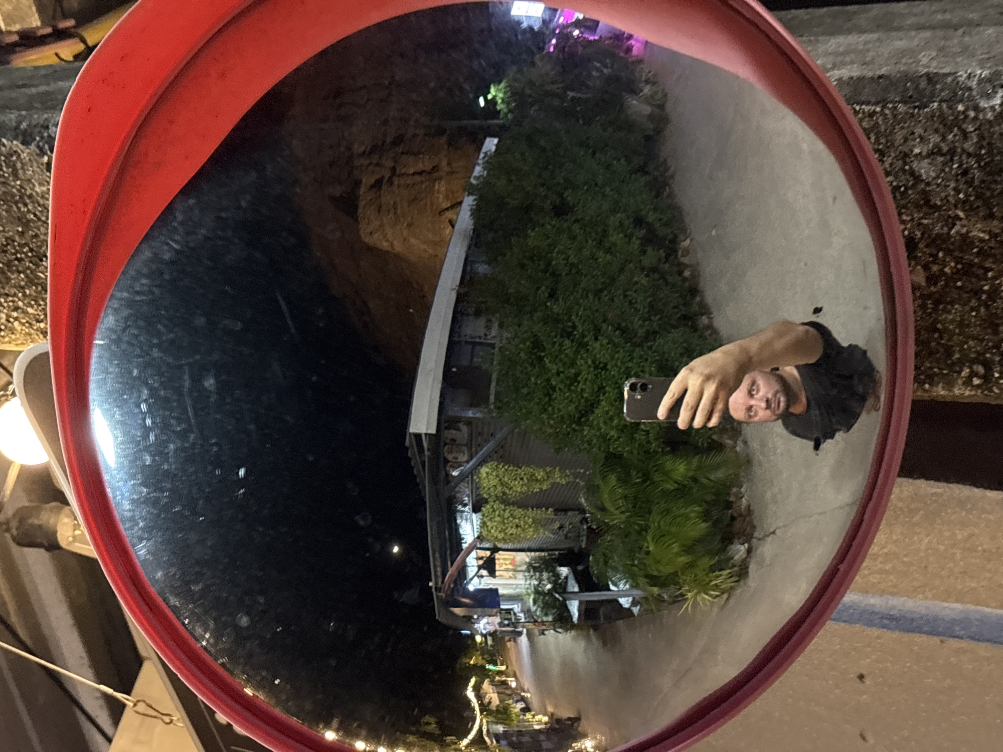 A man takes a selfie in a convex mirror in Baan Bang Rak, Thailand. The mirror, framed in red plastic, reflects a street scene at night. The man holds a phone in his right hand, and his face is visible in the reflection. In the background, there is dense vegetation, a multi-story building with lights, and a dark area possibly indicating a cliff or hill. The ground is paved. A street light is visible in the background. The mirror is attached to a gray metal post.