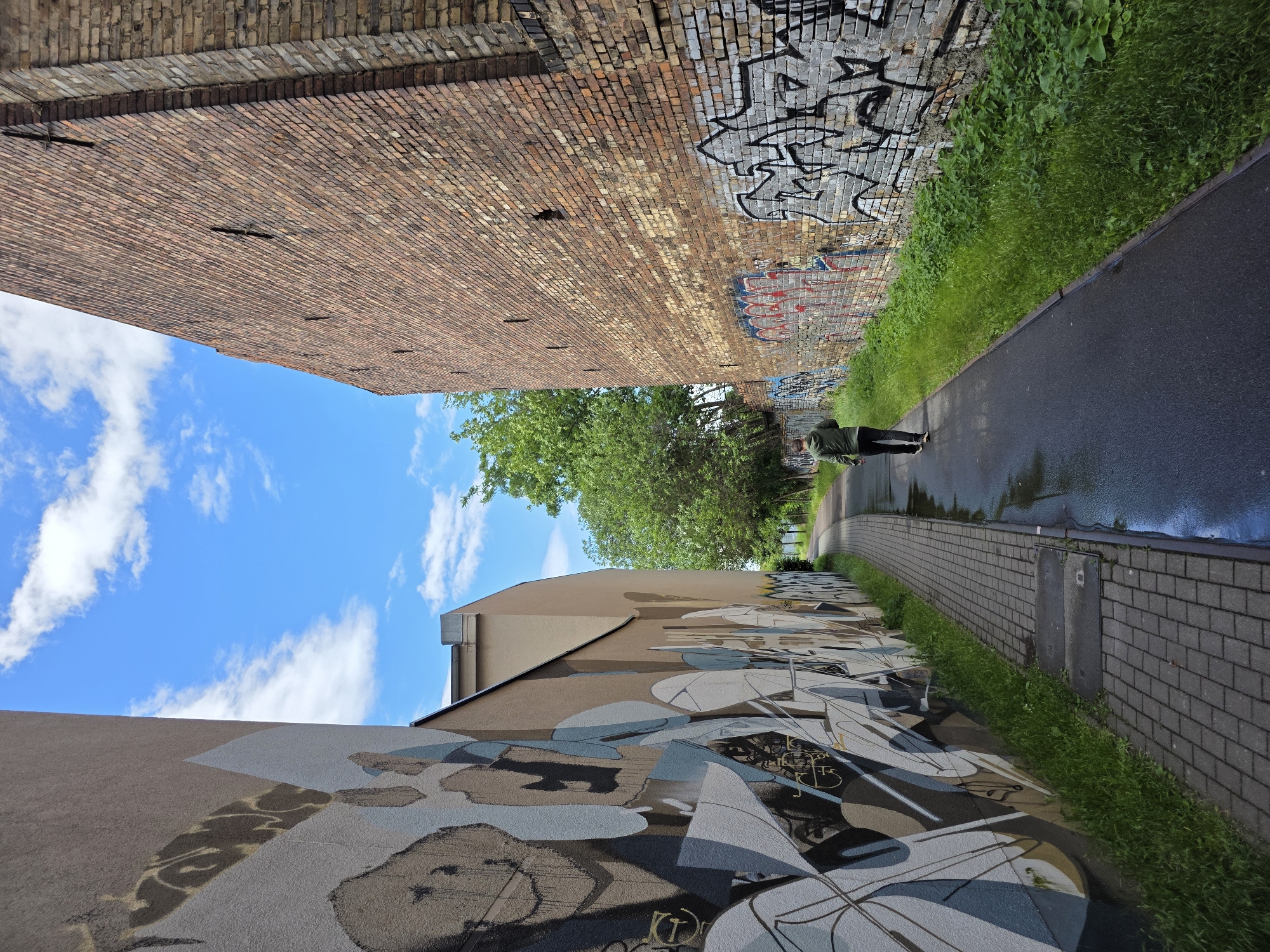 In Halle (Saale), Germany, a person walks along a wet paved path between two buildings. The building on the left is adorned with a large mural. The building on the right is constructed of brick and covered in graffiti. The sky above is blue with scattered white clouds. Green trees are visible in the background.