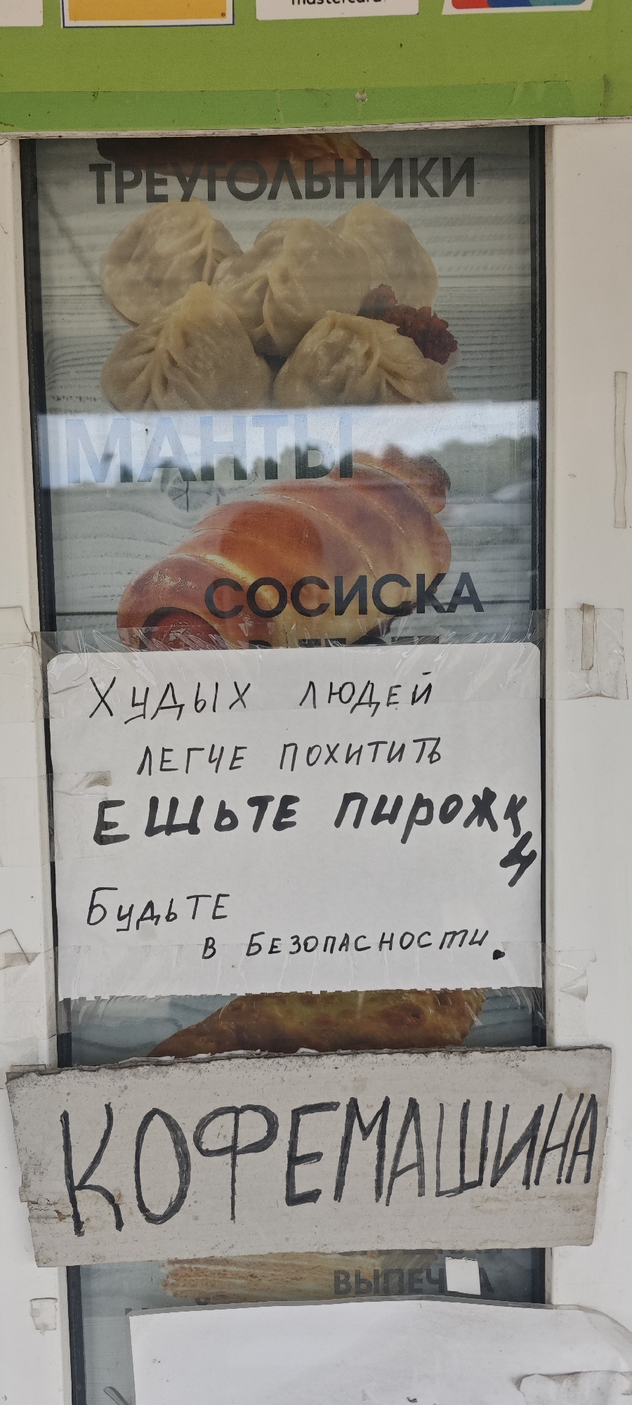 The image shows a window display in Nizhnie Yaki, Russia. The top of the window has a MasterCard logo. Inside the window are advertisements for food items. At the top is a picture of what appears to be dumplings labeled "TRIANGLES". Below that is a picture of a sausage roll labeled "SAUSAGE". Pasted onto the window is a handwritten sign that reads "Thin people are easier to kidnap. Eat pies. Be safe". Below the sign, there is another picture of a pie. At the bottom of the window, there is a sign that says "COFFEE MACHINE". Below that sign, the word "BAKERY" is barely visible.