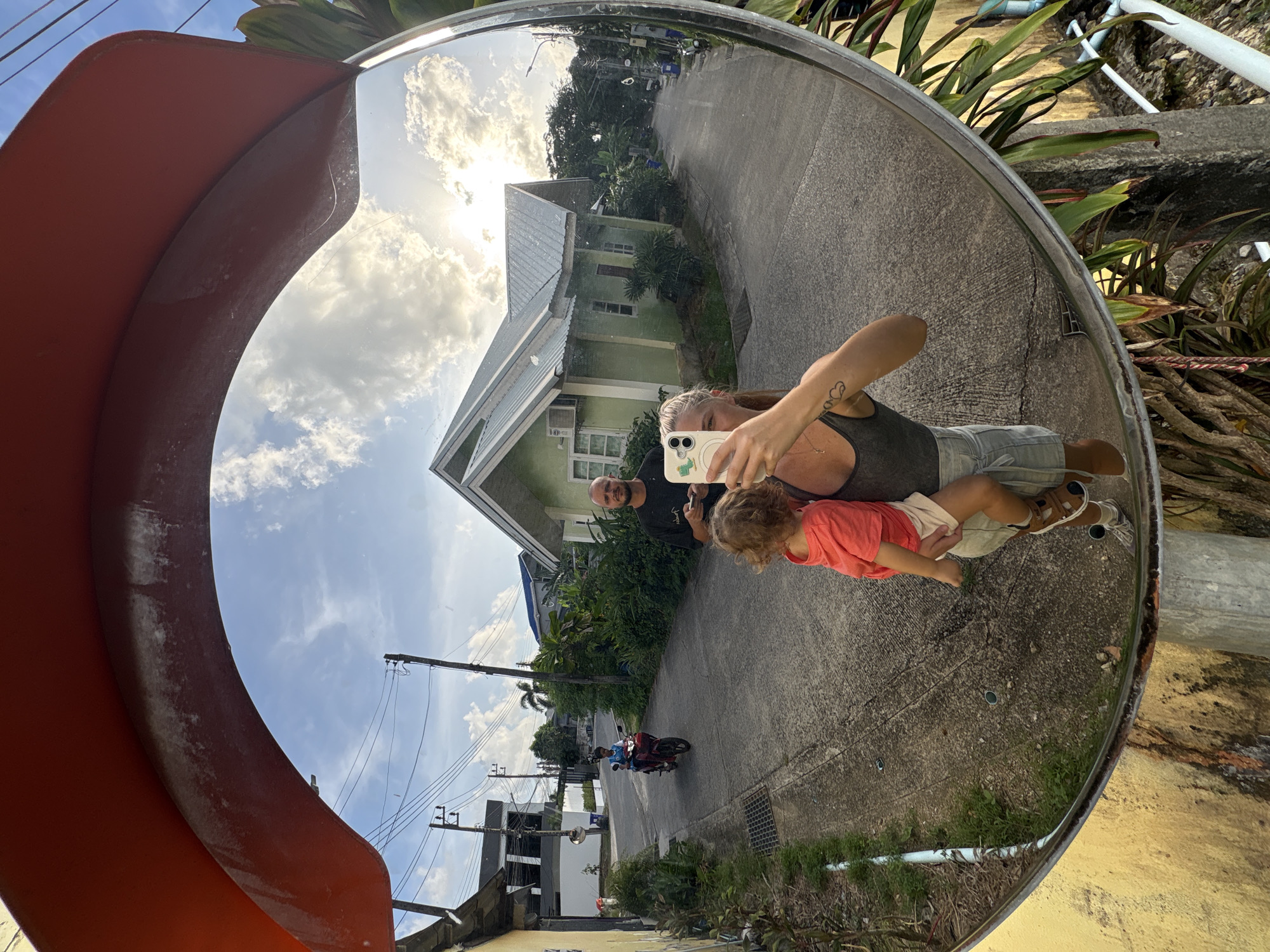 A wide-angle mirror reflects a street scene in Choeng Thale, Thailand, with a woman holding a toddler in the foreground. The woman, with a tattoo on her upper arm, is taking a selfie with her phone. The toddler, wearing an orange t-shirt and beige shorts, is being held in her arms. Behind them, a man with a beard stands looking towards the phone. A young boy rides a red motorcycle down the street. The street is lined with houses and lush vegetation under a cloudy sky. The mirror is mounted on a pole, likely to improve visibility at an intersection.