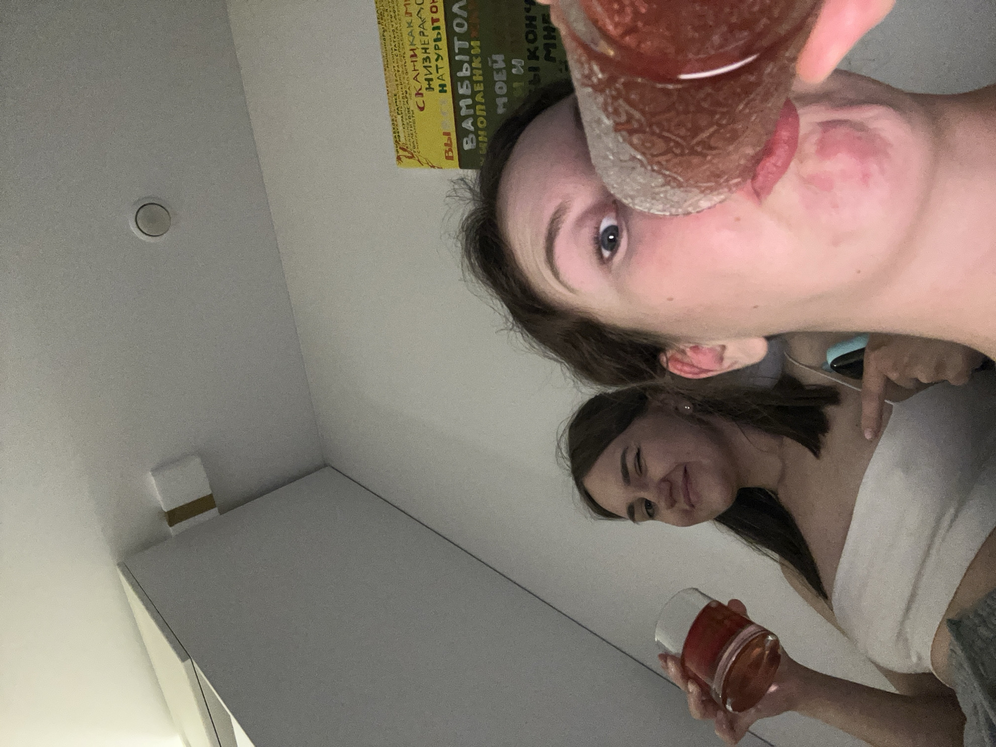 This media file is a low-angle, close-up selfie of two young women in an indoor setting, likely an apartment. One woman, in the foreground and partially cropped, is drinking from a glass of reddish liquid. Her face is turned towards the camera, and her eye is visible. Behind her, another woman is visible, holding a similar glass and looking towards the camera with a slight smile and a wink. Both women appear to be casually dressed.
The background includes a white ceiling with a circular vent or light fixture, and a portion of a wall with what looks like a small white box with a brown band on top, possibly a router or smart home device. In the upper left corner, there is a glimpse of a book with visible text, some of which is in Cyrillic script. The lighting suggests it might be evening or a dimly lit room. The overall mood appears relaxed and informal, with a hint of playfulness from the wink. The presence of books and what might be tech devices suggests a domestic or personal space. The location context of Krasnoyarsk, Russia, is not visually discernible from the image itself.
