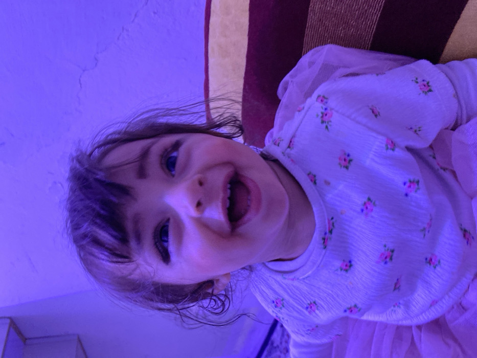 A young child, presumably in the city of Kandahar, Afghanistan, is depicted from the chest up, smiling broadly with an open mouth, looking upward and to the viewer's right. The child has dark hair with bangs, wide blue eyes, pronounced eyelashes, and a dark substance applied around the eyes. They are wearing a white long-sleeved top with small pink floral patterns and a light pink, possibly tulle, skirt. The ambient lighting casts a strong purple hue over the entire scene. In the background, a light-colored wall with minor textural imperfections and cracking is visible. To the right, part of a striped cushion or pillow with reddish-brown and lighter bands is present. A patterned rug or carpet is partially visible on the bottom left. A light-colored, multi-tiered structure is seen on the far left wall.