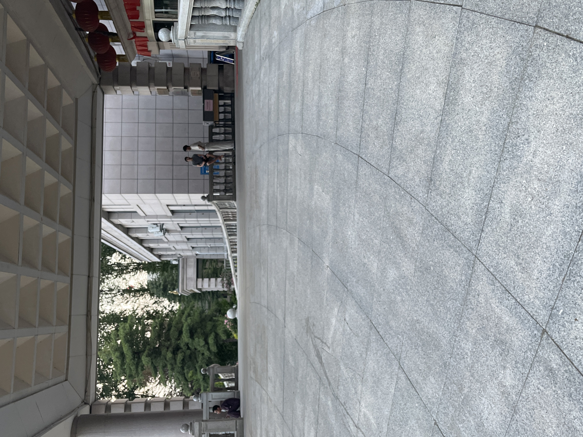 The image depicts a public outdoor space in Changchun City, China. The scene is characterized by the juxtaposition of modern architecture and natural elements. On the left, a building features a facade with a grid-like pattern of square openings, suggesting a covered walkway or ventilation system. Red lanterns hang above this structure, hinting at a festive or cultural context, possibly related to a local event or establishment.
In the background, more of the building is visible, with a light-colored, tiled exterior. A balustrade runs along a raised area, behind which two individuals are standing, facing away from the camera. One person is wearing a light-colored outfit, and the other is in darker clothing. Further back, trees with lush green foliage are visible, indicating a mild to warm climate, and possibly a park or garden adjacent to the building.
The foreground and right side of the image are dominated by a large paved area made of granite tiles. These tiles are laid in a curved pattern, creating a sense of movement and leading the eye into the scene. The texture of the granite suggests it is durable and designed for heavy foot traffic.
A person in a dark suit is partially visible in the lower right corner, crouching down. This individual appears to be interacting with something on the ground or at a lower level.
The lighting suggests it is daytime, likely late afternoon or early evening given the quality of light. The weather appears to be pleasant, with no signs of rain or harsh sunlight. The overall atmosphere is one of everyday activity in an urban environment, with a blend of constructed spaces and natural beauty. No prominent text is visible in the image, except for a possible sign in the background that is too small to read.