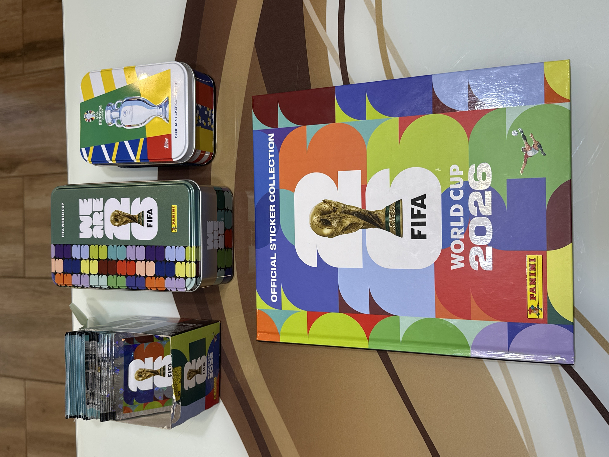 The image shows three collectible tins and an open sticker album on a table. One tin is green and features the FIFA World Cup 2026 logo and the FIFA World Cup trophy. Another tin is also green with a colorful geometric pattern and the words "WE ARE ONE". The third tin is white with a design of the Euro 2024 trophy and is labeled "OFFICIAL STICKER COLLECTION". The sticker album is open, revealing a colorful cover with the FIFA World Cup 2026 logo and the trophy. The setting appears to be indoors, on a table with a patterned surface. There are no people present in the image. The image does not contain any information about the city of Prisovo, Bulgaria.