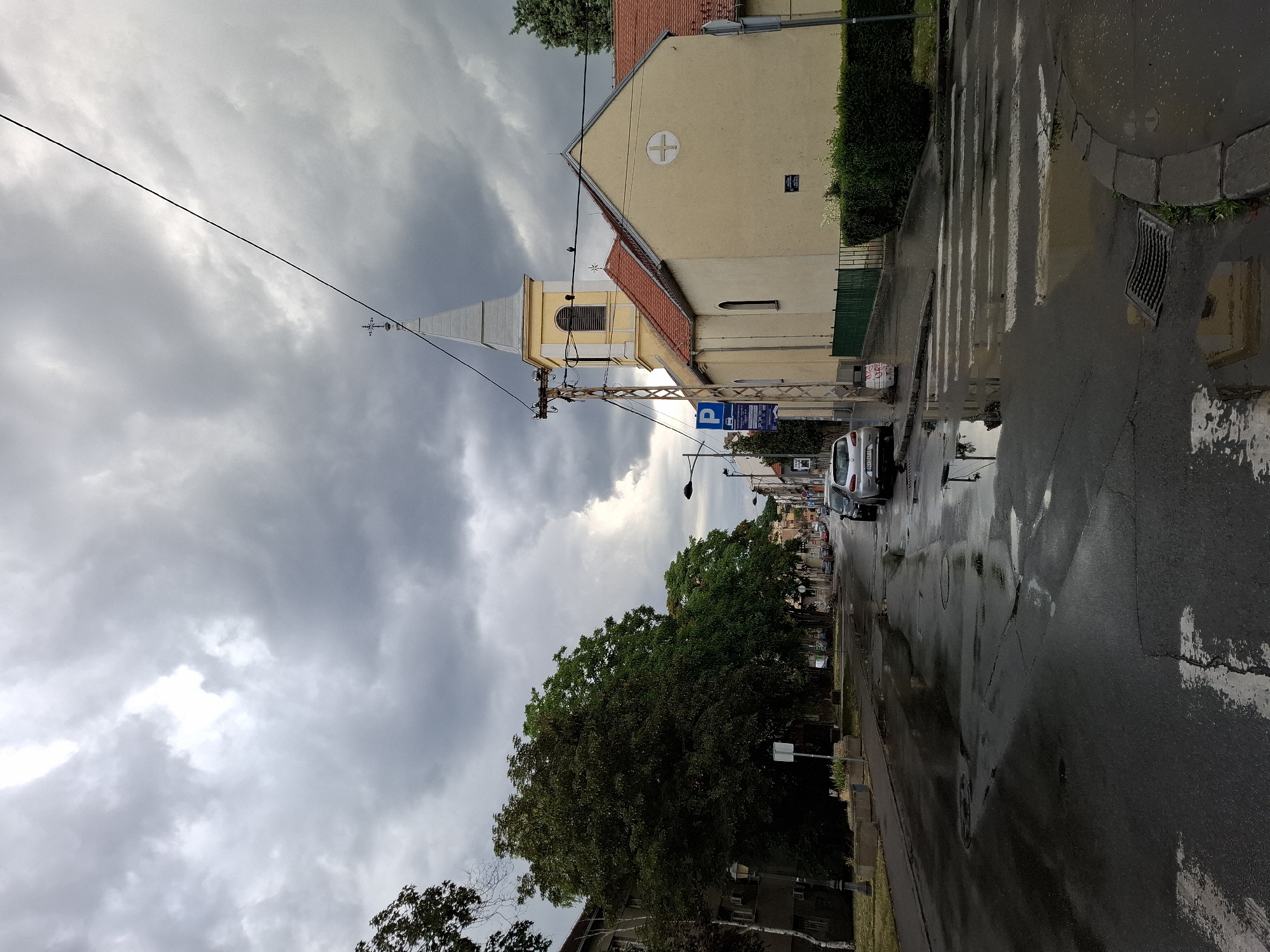The image depicts a street scene in Zemun Urban Municipality, Serbia, under a heavy, overcast sky, suggesting recent or impending rain. The street is wet and reflects the surrounding environment, with puddles evident on the asphalt.
On the right side of the street, a pale yellow building with a red-tiled roof is prominent. It has a steeple topped with a cross, indicating it is likely a church. The building features a circular emblem with a cross on its side. A green hedge runs along the front of the building.
A pedestrian crossing is visible in the foreground, with white stripes partially submerged in water. A metal drain grate is located on the curb.
To the left of the street, there are trees with lush green foliage, some of which overhang the pavement. A building with several windows can be seen behind the trees. Streetlights and lampposts are also present along the sidewalk.
Several cars are parked and driving on the street. A silver car is parked on the right side of the road, and others are visible further down the street.
A blue and white sign with a "P" symbol, likely indicating parking, is mounted on a pole near the church. Another sign is visible on a pole further down the street.
The overall atmosphere of the image is somewhat somber due to the dark clouds and wet conditions, yet it captures a typical urban street scene.
