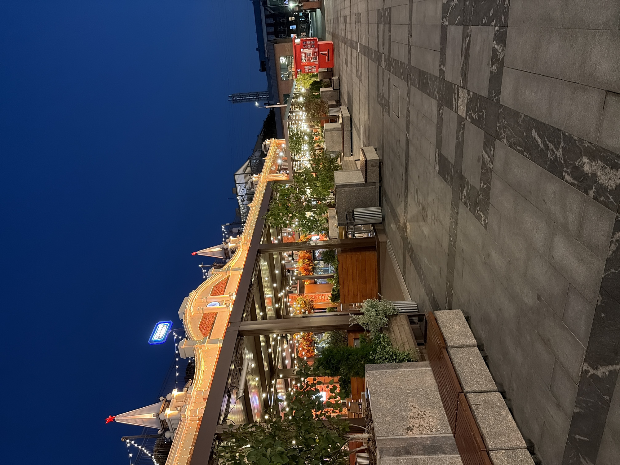 The image depicts an outdoor dining area at night, likely part of a restaurant or cafe, located in Moscow, Russia. The architecture features a long, illuminated building with a red-brick facade and a covered patio area adorned with numerous string lights. Several trees in planters, along with bushes, are visible in front of the dining area. The ground is paved with large grey tiles, some arranged in a decorative pattern with darker grey marble inlays. Benches, also made of grey stone and wood, are placed along the pathway. In the background, a bright red kiosk stands on the right side of the paved area, and further in the distance, other buildings and a street lamp are visible against the dark blue night sky. There are no people in the frame. The overall impression is of a well-lit, urban outdoor space suitable for evening leisure.