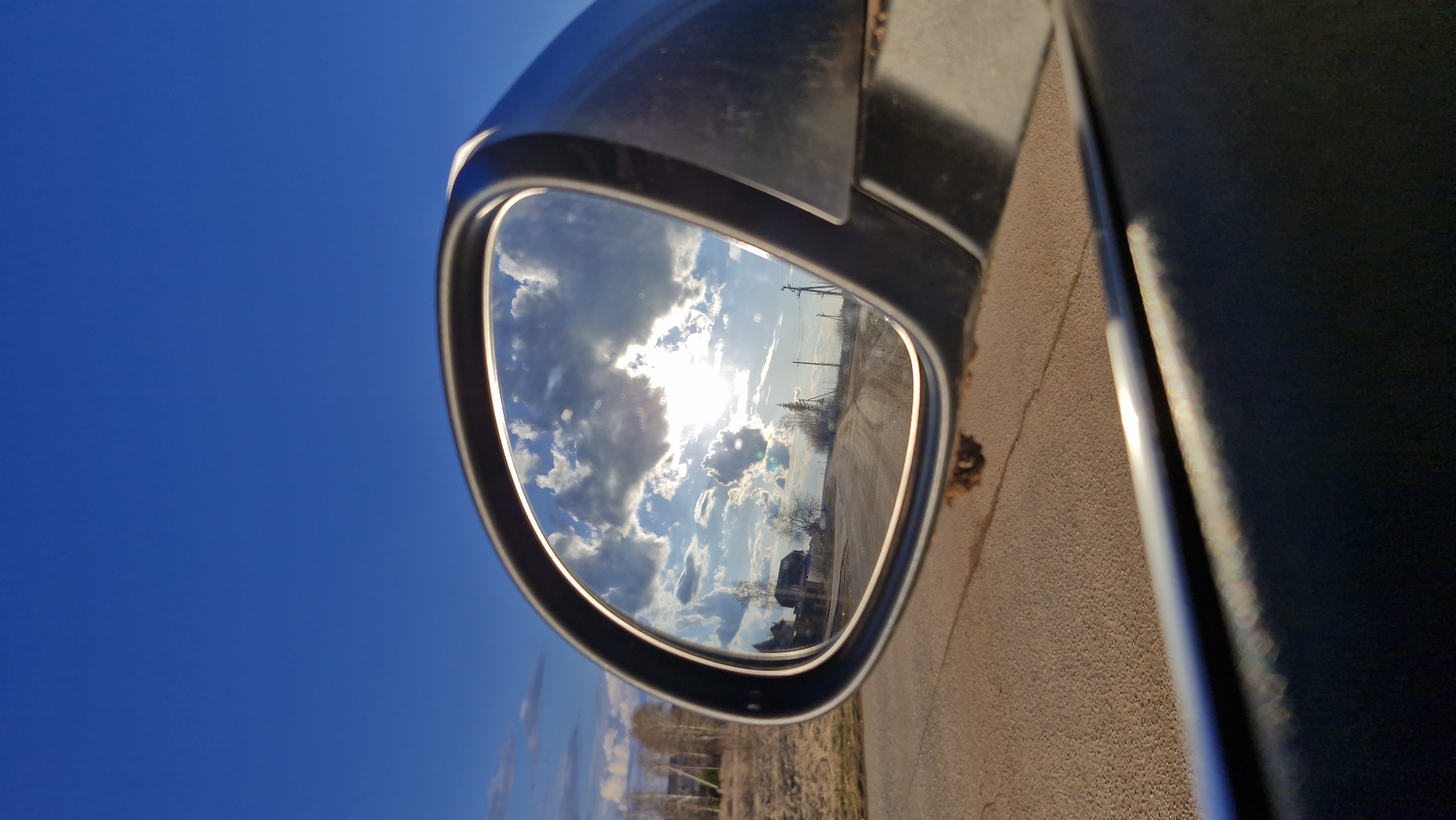 A slightly tilted, outdoor shot centers on the side mirror of a dark-colored vehicle, likely a car. The image is rotated, presenting the horizon diagonally within the mirror's reflection.
The primary subject is the car's side mirror, framed by the dark body of the vehicle and a vibrant, deep blue sky to its left. Within the mirror's reflective surface, a dynamic sky scene unfolds: bright sunlight bursts from behind a cluster of fluffy white and gray clouds, creating a strong glare in the upper-center. Scattered clouds fill the rest of the blue sky visible in the reflection.
Below the sky in the mirror, a rural landscape is depicted. A light brown, possibly dirt or gravel, road stretches into the distance on the right. Several tall power poles with visible lines run along the side of this road, and sparse, bare trees or bushes are scattered across the landscape. A small building or house can be vaguely discerned in the mid-ground, slightly to the left of the road.
Outside the mirror, to the right, a dark, reflective surface of the car's body is visible, partially shadowed. A strip of light brown ground, similar in texture to the road in the reflection, runs along the bottom-right edge of the frame. The clear, deep blue sky, devoid of clouds, occupies the upper-left portion of the image, contrasting with the cloud-filled reflection.
The strong sun glare and the angle of the light suggest either early morning or late afternoon. The bare trees and muted ground colors hint at late autumn, winter, or early spring. The setting appears to be a rural or semi-rural area, consistent with Otchor, Russia. There is no visible text in the image.