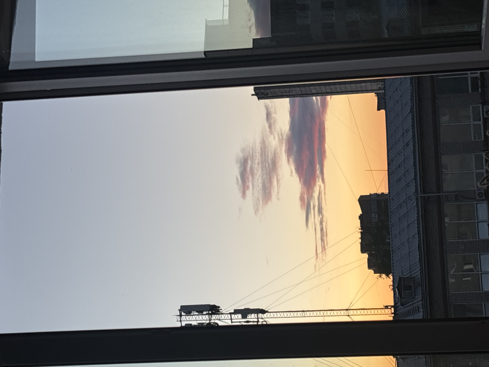 The image shows a view through a window of a city skyline at sunset. The sky is filled with warm orange and pink hues from the setting sun, with some scattered clouds. In the foreground, a metal cell tower is visible on the left, with several antennas attached. Several buildings are visible in the middle ground, including a multi-story building with a flat roof and several windows, and a taller, darker building to its right. The lower part of the image shows the roof of a brick building with a corrugated metal roof and several windows. The scene is framed by the dark edges of the window frame. The overall impression is of a typical urban environment during twilight.