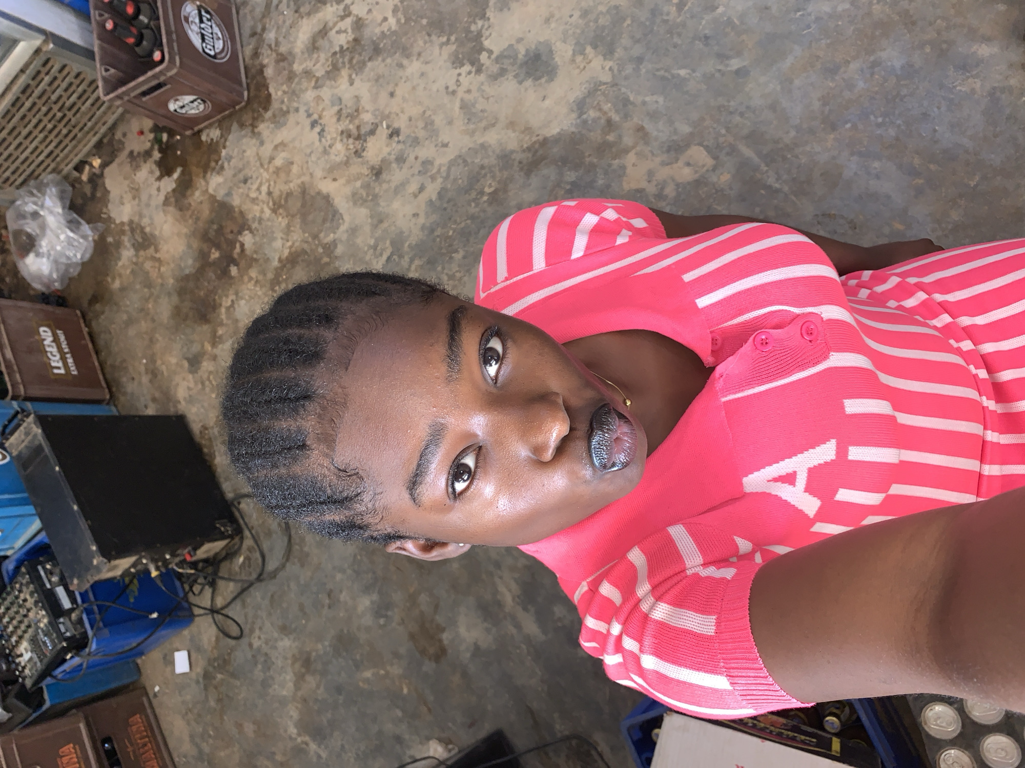 A woman with braided hair and dark skin, wearing a pink and white striped shirt, is lying on a concrete floor. Her face is directed towards the camera with her mouth slightly open. Several boxes, some containing bottles, are scattered around the room. A black electronic audio mixer and other electronic equipment with wires are located to the left of the woman. The scene is illuminated by natural light. This appears to be an informal setting, possibly a warehouse or storage area within the city of New Stadium, Nigeria.