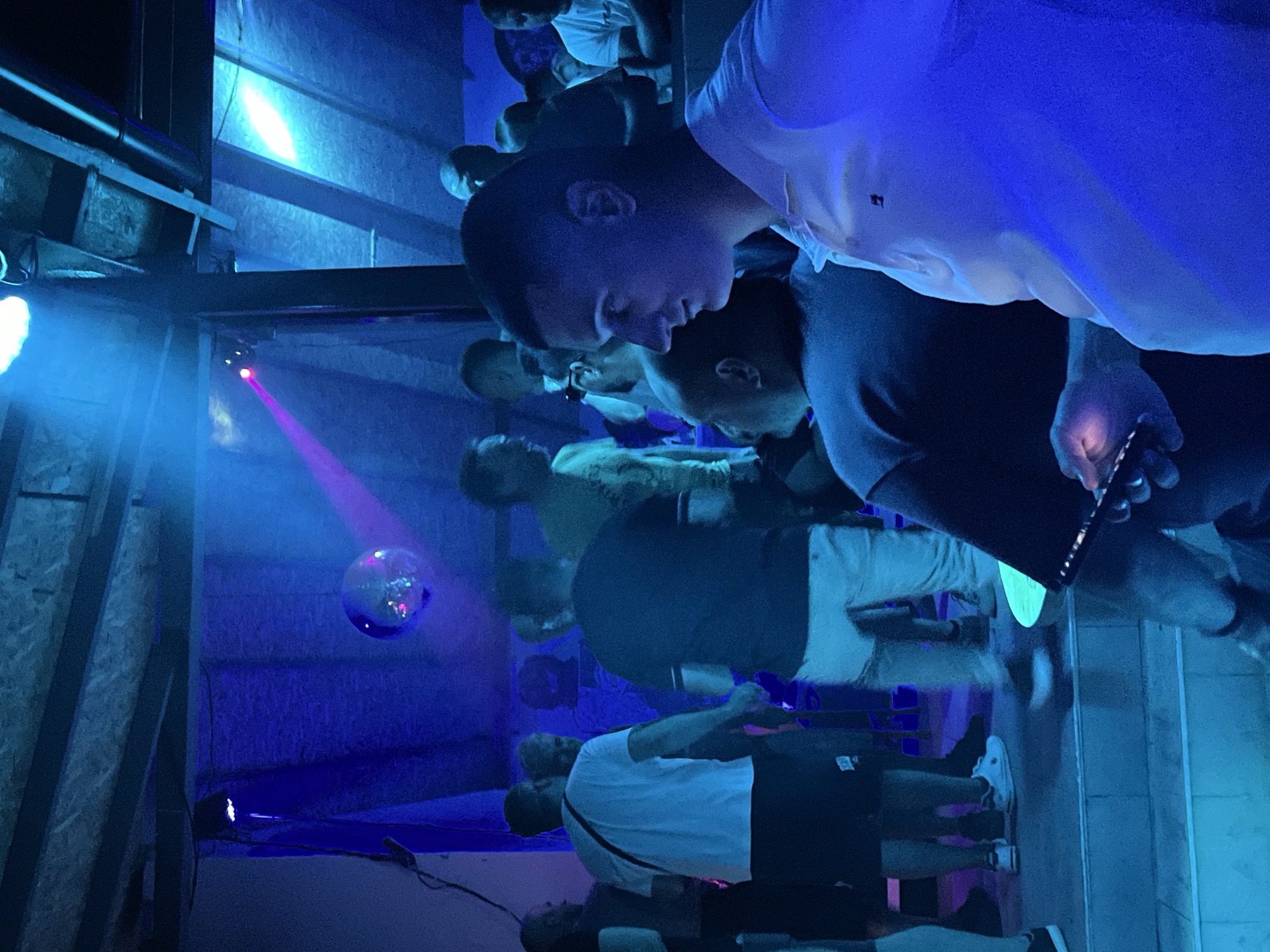 The image depicts an indoor gathering, possibly a club or bar, illuminated by blue and pink stage lighting. In the foreground, a man in a white polo shirt is looking to the side, holding a phone. Behind him, several other individuals are visible, some standing and some seated. A disco ball hangs from the ceiling, reflecting the lights. The environment appears to be rustic, with exposed wooden beams and draped fabric. The scene suggests a social event or party. The location is confirmed to be in Herceg Novi, Montenegro.