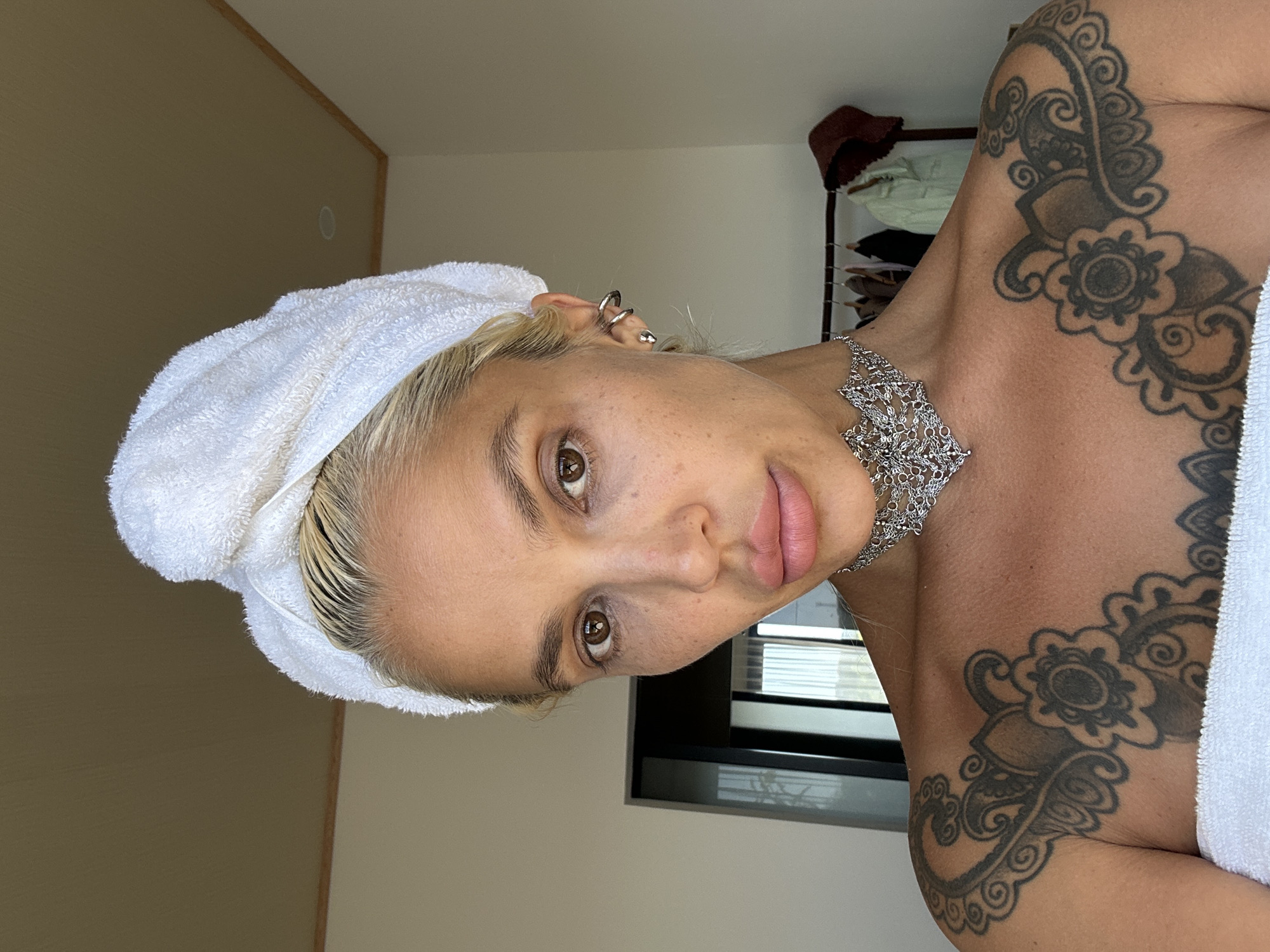An adult female is depicted in a close-up, chest-up portrait, inside a room in Kuta Selatan, Indonesia. She has platinum blonde hair secured in a white terry cloth towel wrapped in a turban style. Her face is directed towards the camera, revealing brown eyes, full lips with light pink lipstick, and visible skin texture with scattered freckles. She wears a wide, ornate silver choker necklace with intricate patterns and multiple silver earrings in her right ear, including two cartilage hoops and a lobe stud. Extensive black ink tattoos, featuring floral, mandala, and swirling designs, cover her upper chest and shoulders. A white towel is wrapped around her upper body, covering her chest. The background consists of light-colored walls and a light brown ceiling with wooden trim. On the left, a dark-framed glass doorway or window is partially visible. On the right, a wooden clothing rack holds several garments, including a light green top and darker items, along with a reddish-brown hat.