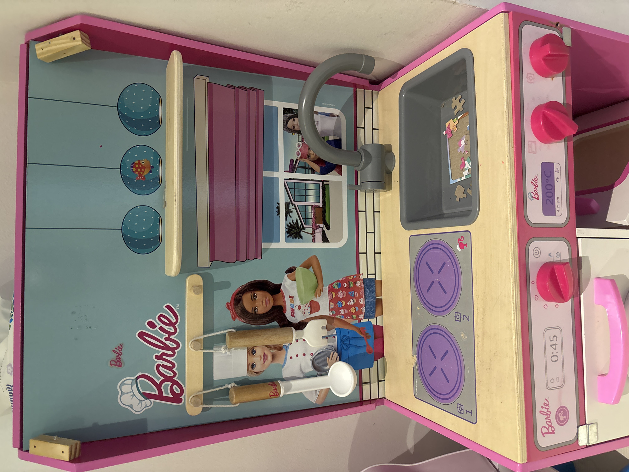 A children's pink and teal wooden toy kitchen, designed to resemble a Barbie-themed play set, is the subject of this image. The kitchen features two purple stovetop burners with stylized cross markings, a gray sink with a faucet, and a control panel with a digital display showing "0:45". Above the stovetop and sink, a backdrop depicts two Barbie dolls, one in a chef's hat and the other in an apron decorated with cupcakes. Hanging utensils, including a spoon and a spatula, are suspended above the dolls. The upper section of the kitchen has a shelf with decorative spherical lamps, one featuring a small fish motif. In the background, behind a window depicted on the backdrop, are two other Barbie figures. The toy kitchen is positioned against a white wall. The image does not provide information to determine its location in Dubai, United Arab Emirates.