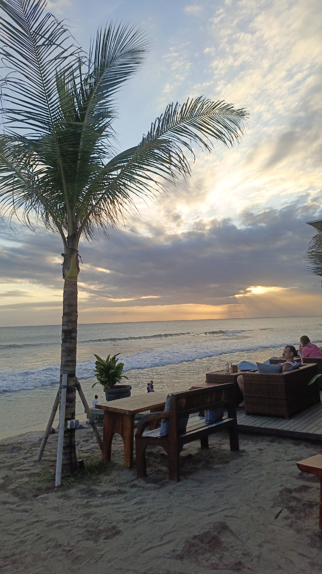 The image depicts a beach scene in North Kuta, Indonesia at sunset. A palm tree stands on the left, supported by wooden poles. A wooden table and bench sit on the sand. Two people sit on wicker furniture on a slightly elevated wooden platform to the right, observing the ocean. The sun is setting, with rays of light streaming through the clouds. Waves are visible in the ocean. Two figures are visible in the water. A potted plant sits near the palm tree.