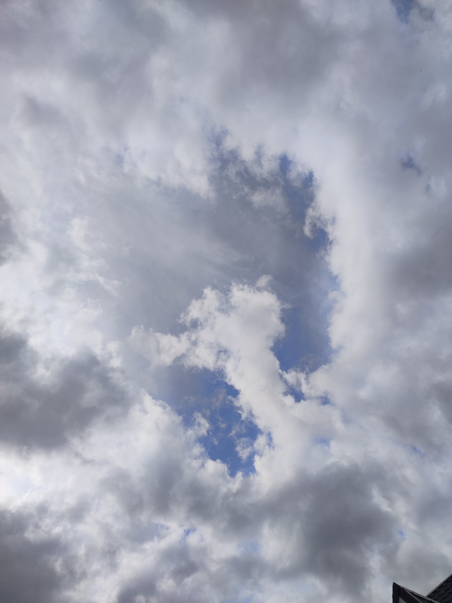 The image displays an overcast sky filled with numerous clouds, with a small portion of a building roof visible in the bottom right corner. The sky is predominantly covered by a mix of dense, textured clouds ranging in color from bright white to various shades of grey, indicating varying thickness and light exposure. Scattered patches of clear blue sky are visible through the cloud cover, particularly towards the center and upper left, suggesting breaks in the clouds.
The clouds exhibit different formations, with some appearing soft and wispy, while others are more voluminous and solid. The lighting suggests it is daytime. In the bottom right corner, a dark, presumably tiled roof with a light-colored trim is partially visible, indicating the photograph was taken looking upwards from near a structure.
There are no people, specific activities, or discernible objects other than the clouds and the roof fragment. No visible text is present. The scene depicts a typical mixed-weather sky in Fokino, Russia.