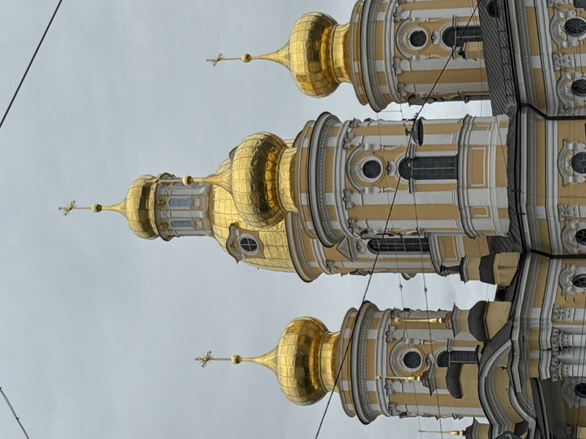 The image shows an upward view of the Smolny Cathedral in Saint Petersburg, Russia. The cathedral features multiple golden domes with crosses at the top. The building's exterior is decorated with white and yellow vertical stripes, and round windows. Overhead power lines are visible against the overcast sky.