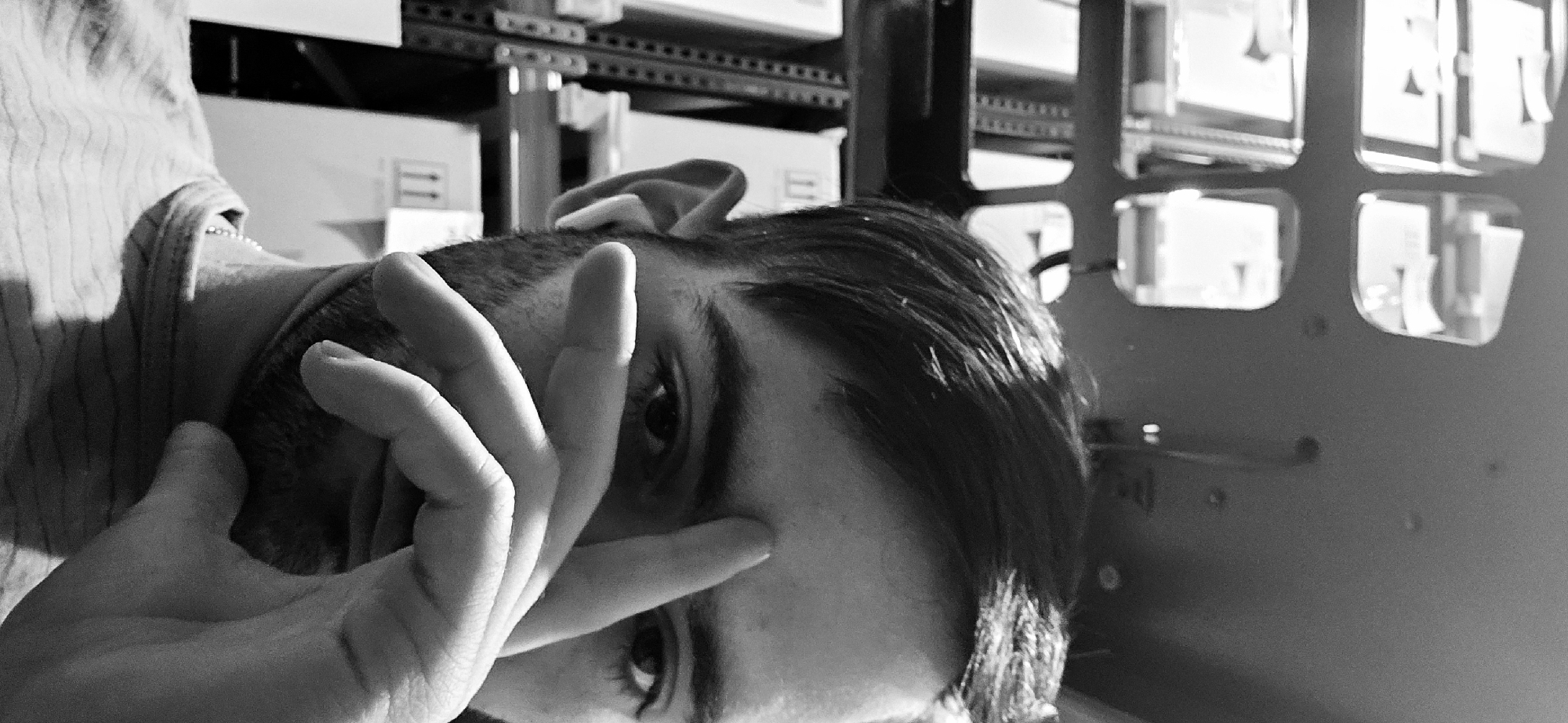 A black and white image, tilted on its side, captures a person looking towards the viewer while partially obscuring their face with their right hand. The person's left eye is clearly visible, making direct eye contact, with a portion of the right eye also visible beneath the fingers. They have short, dark hair, styled to the side, and a faint stubble. A striped pattern is visible on the shirt or garment worn by the person, on the left side of the frame. A thin, beaded chain or necklace is visible around their neck.
The background suggests an indoor environment, likely an industrial, office, or storage area. Visible behind the person are rows of shelves or racks holding various boxes and containers. Some of these boxes show symbols resembling "11" or a pair of right-pointing arrows. On the right side of the image, a dark structure, possibly part of a server rack or cabinet, features several square and rectangular cutouts through which blurred, bright objects are visible. The strong lighting in the scene casts noticeable shadows, adding depth and contrast to the composition. No specific text is clearly legible other than the "11" or arrow symbols on the boxes.