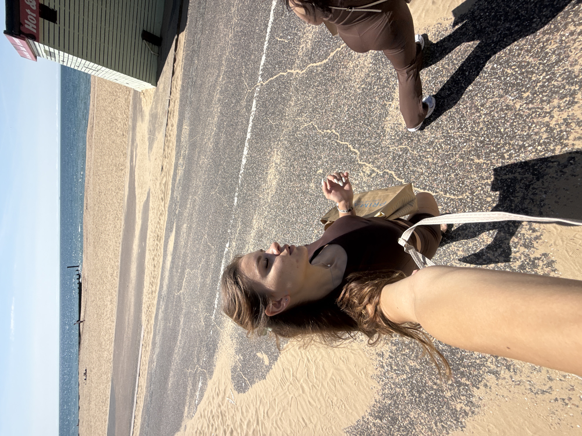 The image depicts a bright, sunny day at the beach in Bournemouth, United Kingdom. In the foreground, a young woman with long brown hair, wearing a brown one-piece swimsuit, is seen from a high-angle perspective, holding a shopping bag. To her right, another person, also wearing brown athletic wear, is partially visible, in motion. Sand has accumulated on the asphalt pathway, creating a contrast with the dark grey surface. The background shows a sandy beach, the blue ocean, and a clear sky. A small, light-green beach hut with a red roof, labeled "Hot &," is situated on the right side of the frame. The overall scene suggests a casual outing by the sea.