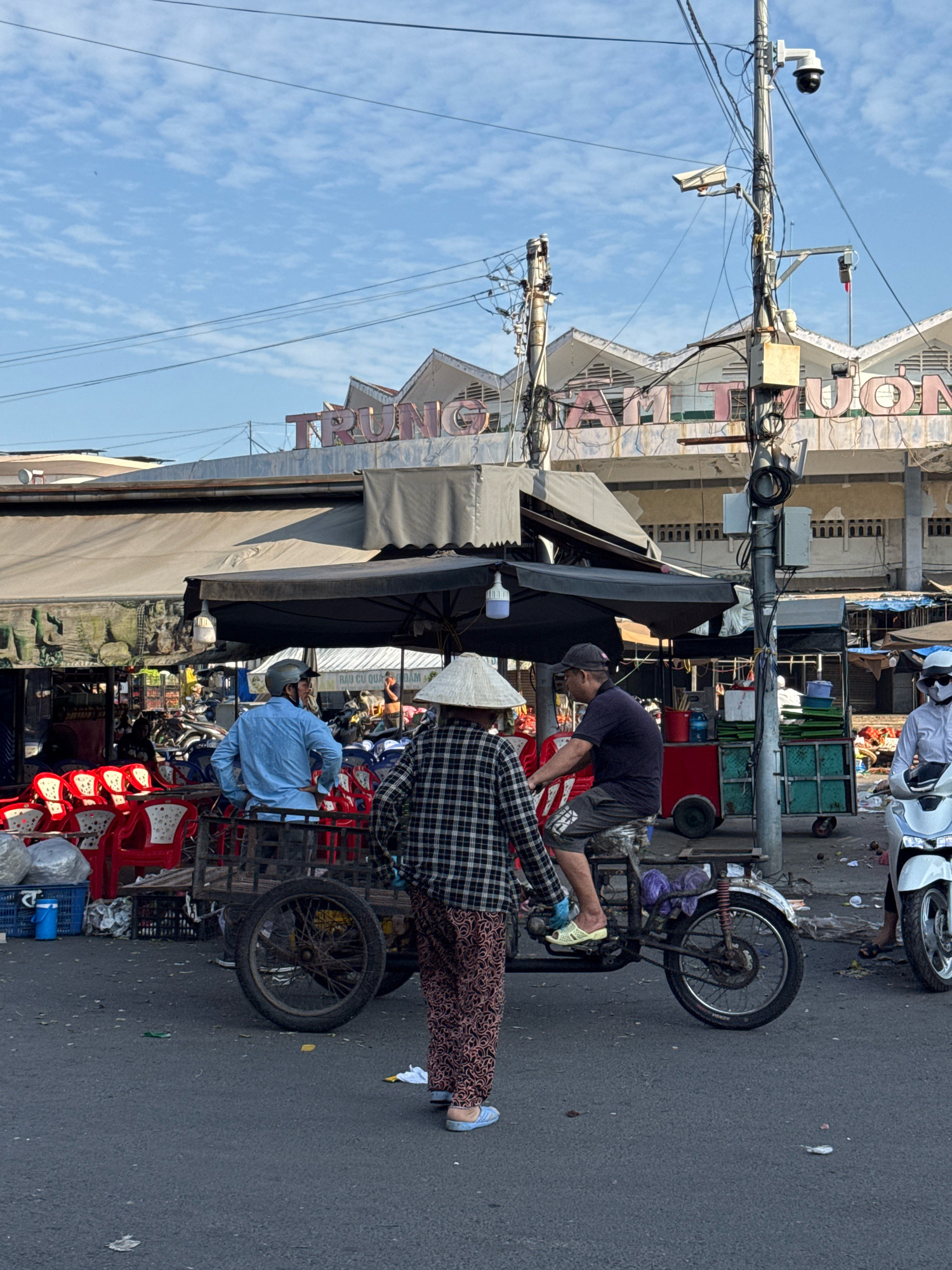 In Nha Trang, Vietnam, an outdoor market scene unfolds under a clear, blue sky with scattered clouds. A central focus is a man on a modified tricycle-like vehicle, wearing a dark t-shirt and shorts, with a cap on his head. To his left, a person in a blue shirt and helmet stands by a cart filled with red chairs. In the foreground, facing away from the camera, is a person in a checkered shirt and patterned pants, wearing a conical hat. The background reveals a building with "TRUNG TAM THUONG" signage and a marketplace with stalls and more vehicles, including a white scooter on the right. The ground is paved asphalt, littered with some debris. The overall impression is one of everyday activity in a bustling Vietnamese market.