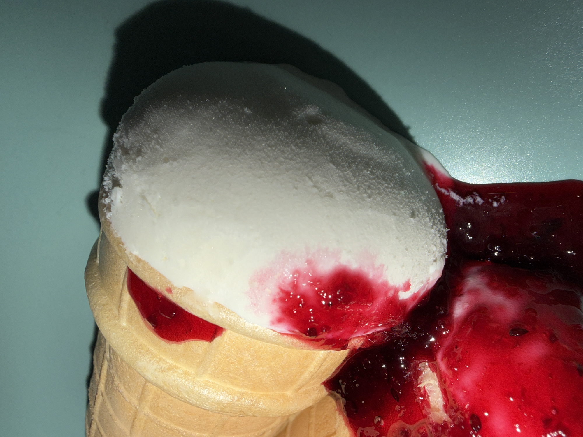 The image presents a close-up, high-angle view of a melting ice cream cone with berry sauce. A large scoop of white ice cream, appearing cold and slightly frosted on its surface, sits within a light brown waffle cone. Vivid, glossy red berry sauce or jam is abundantly poured over the ice cream and cone, and is visibly pooling on the light blue-green surface where the cone rests. Darker berry pieces are discernible within the rich red sauce. Streaks of pink where the sauce is melting into the white ice cream indicate its melting state. The waffle cone exhibits its characteristic textured pattern. A distinct, dark shadow is cast behind and to the left of the ice cream cone, suggesting illumination from the right or front. The scene captures the immediate aftermath of a dessert being served, with the ice cream rapidly melting. No people are visible, nor is any text present. The given location context is Chaikovsky, Russia, though this is not visually evident in the image itself.