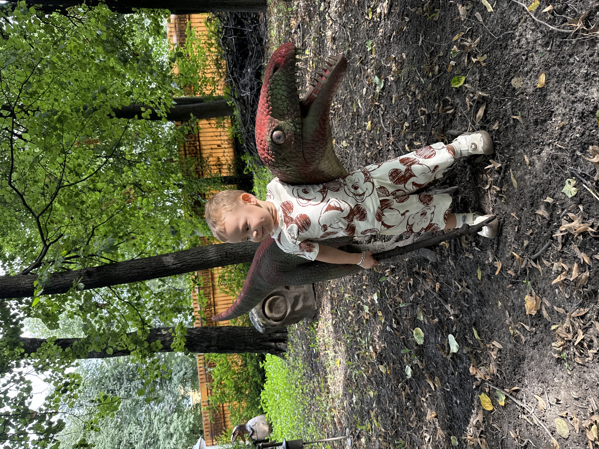 A young child stands to the right of a dinosaur statue in a park setting. The child, wearing a patterned outfit and white sneakers, is looking to the left. The dinosaur statue is positioned in the foreground, with its head and neck visible. In the background, there are trees and a wooden fence. The ground is covered with dirt, leaves, and scattered branches.
