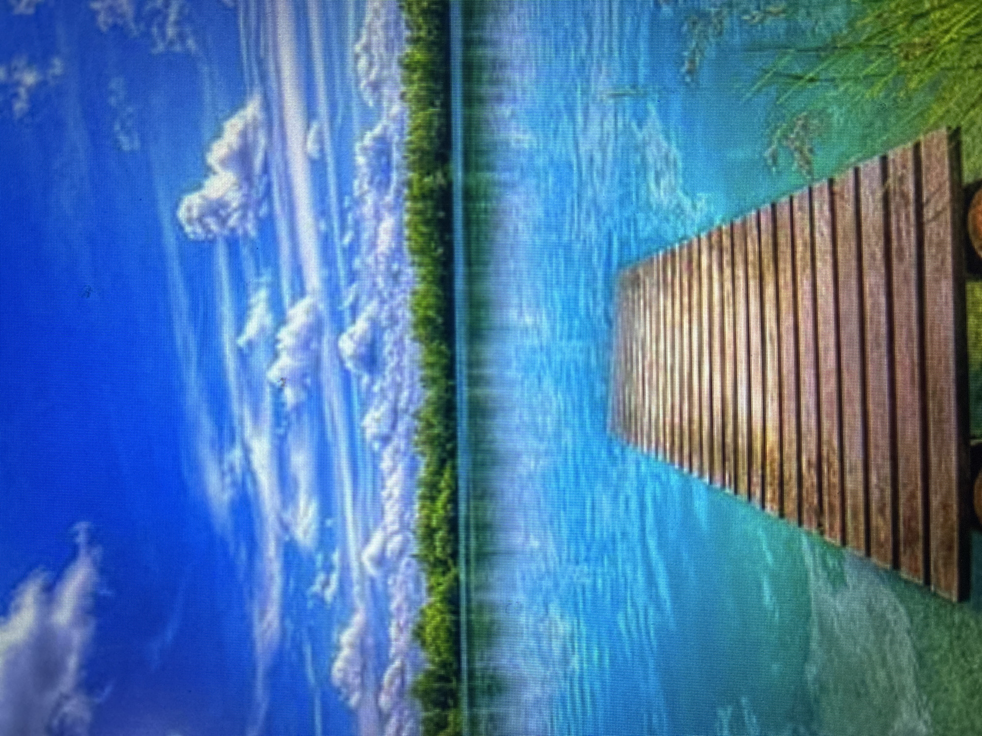 This image presents a serene natural landscape, most likely a lake or a large river, under a bright, cloud-dotted sky. The scene is oriented as if looking across the water towards a distant shore.
In the foreground, a wooden pier extends into the clear, blue-green water from the right side of the frame. The pier is constructed from multiple parallel wooden planks, which show signs of weathering, suggesting an older structure. A small, round, dark object is visible at the very end of the pier where it meets the shore on the right, possibly a wheel or a part of its support system.
The water itself is calm, reflecting the vibrant blue of the sky and the green hues of the nearby land. Subtle textures and ripples are visible on its surface. Beyond the body of water, a dense treeline or forest forms a continuous green band across the horizon, separating the water from the sky.
Above the treeline, the sky is a brilliant blue, populated by numerous white, fluffy cumulus clouds that appear stretched or streaked, possibly due to either artistic rendering or motion. The bright lighting indicates it is daytime, likely a sunny day, creating a peaceful and tranquil atmosphere.
No people or specific activities are depicted, focusing solely on the natural environment. There is no visible text in the image. The image quality, particularly visible in the crops, suggests it is a photograph of a digital screen, with a noticeable pixel grid, and the original image is rotated 90 degrees clockwise. Given the context, this tranquil scene is located near Asenovgrad, Bulgaria.