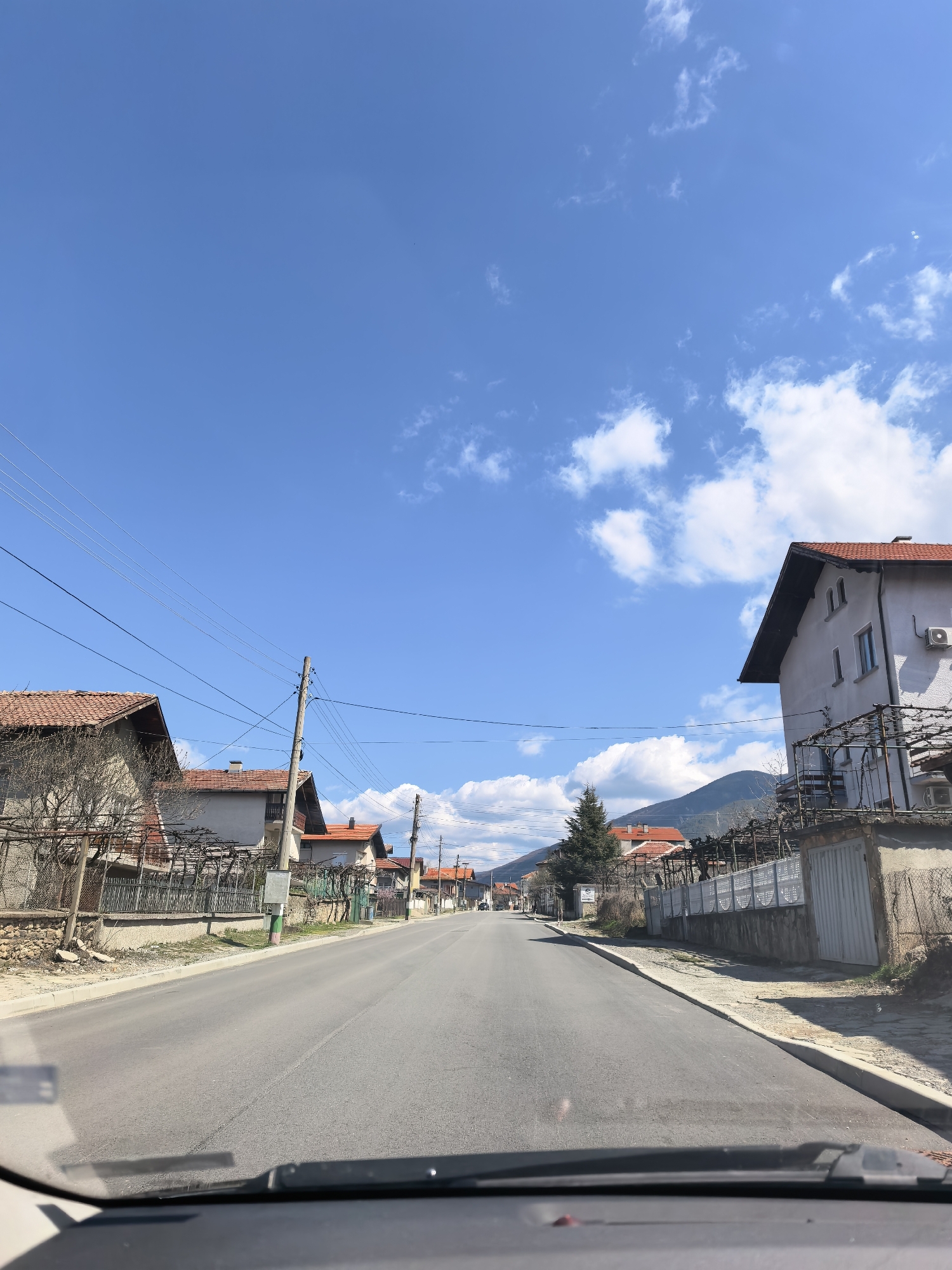The image is taken from inside a vehicle, likely a car, looking out onto a residential street in the city of Unknown, Unknown. The street is paved and stretches straight ahead, with houses lining both sides. The houses are a mix of single and multi-story buildings, with some having red tile roofs. Utility poles and wires run along the left side of the street. The sky is blue with scattered white clouds. In the distance, a mountain range is visible. The overall scene suggests a quiet, rural or suburban neighborhood.
