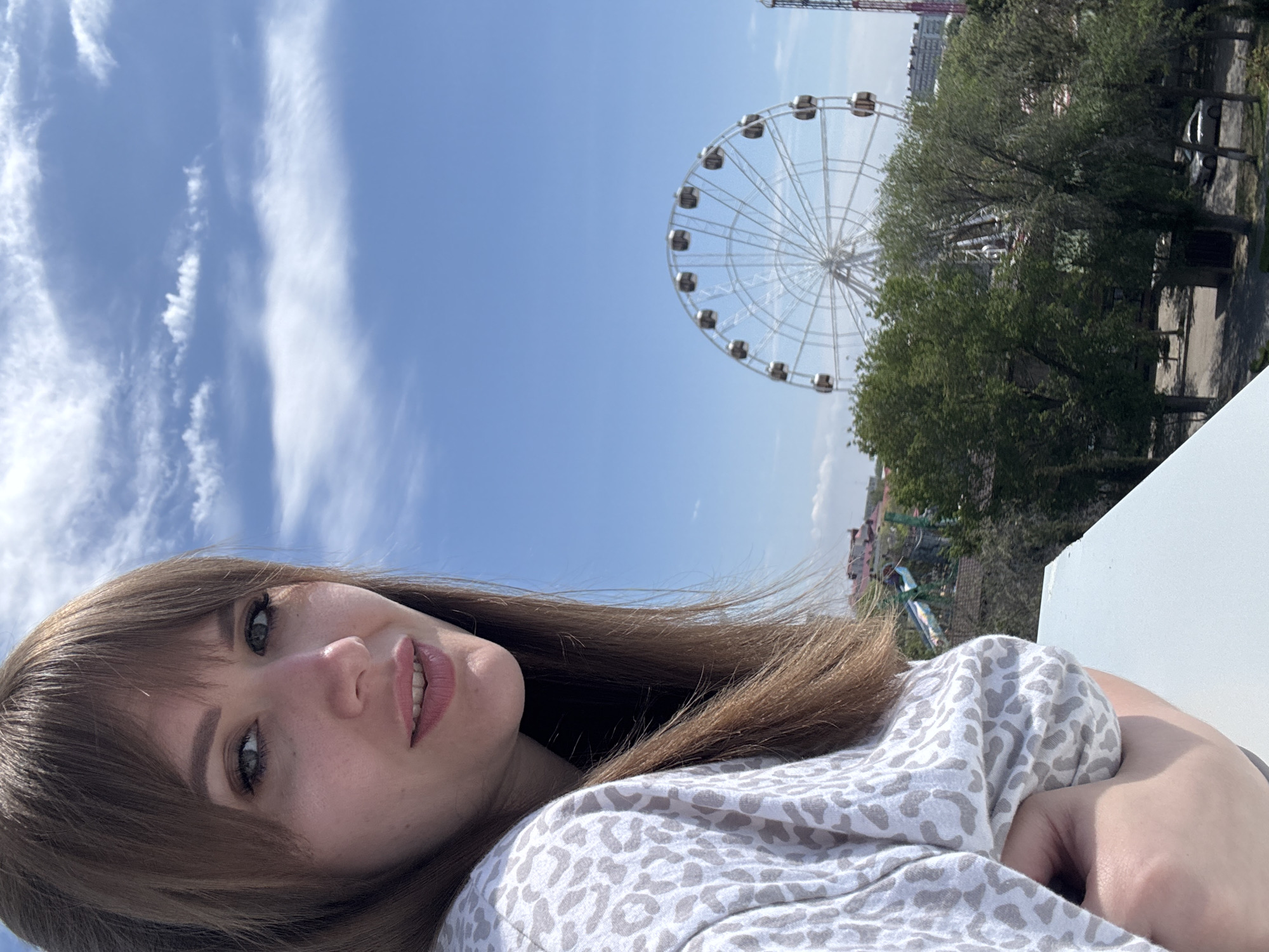 A medium shot shows a woman in the foreground and a Ferris wheel and trees in the background. The woman has long brown hair with bangs. She has fair skin, and is wearing a white and gray spotted top. Her arms are crossed in front of her. Behind her to the upper left, is a blue sky with white clouds. To the upper right is a Ferris wheel with many white seats, and beyond it a building. Behind the Ferris wheel is dense green foliage. It appears the picture was taken in Yevpatoriya, Ukraine, due to the Ferris wheel’s location in the city.