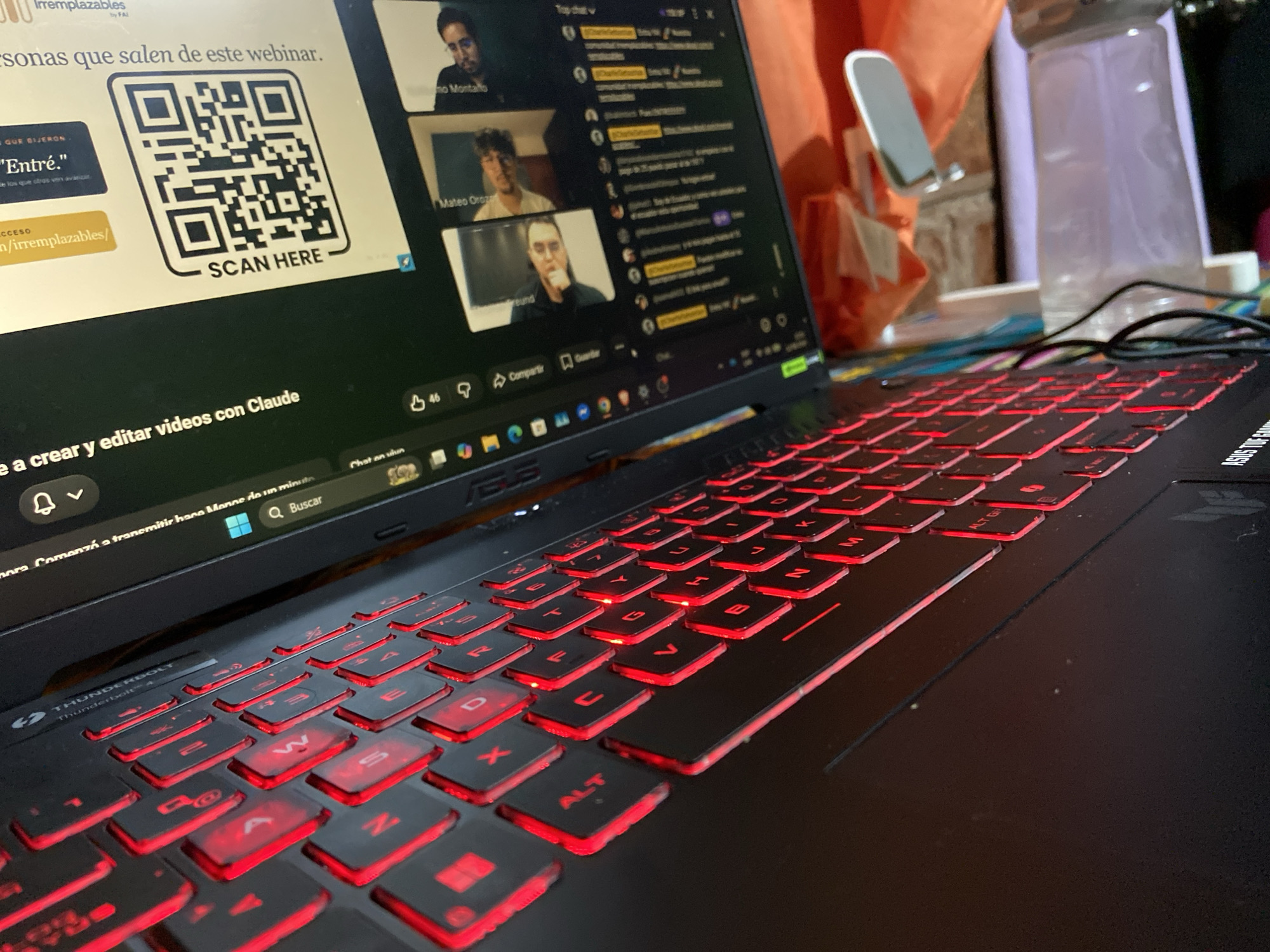 The image shows a close-up of a laptop with a black keyboard illuminated by red backlighting. The laptop screen displays a video conferencing interface with several participants visible. On the left side of the screen, a QR code is displayed with the text "SCAN HERE". The overall environment appears to be indoors, possibly a home office, with some blurry background elements including an orange cloth, a clear bottle, and other desk items. There are no specific visual cues to suggest a location in Pelileo, Ecuador. The image focuses on the laptop and its contents, highlighting the technological aspect of a virtual meeting.