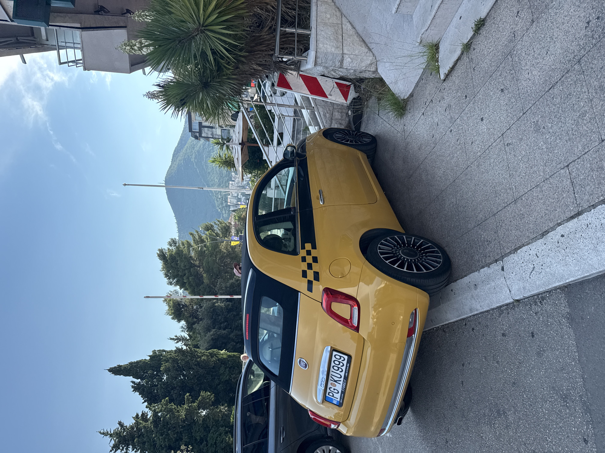 The image depicts a yellow Fiat 500 car parked on a sidewalk next to a street. The car has black trim and a black convertible roof. A checkered pattern is visible on the rear pillar of the car, typical of a taxi. The license plate reads "PG KU999". In the background, there are trees, buildings, and a hill. A striped barrier is also visible near the car. The sky is clear and blue, suggesting a sunny day. The scene appears to be in an urban or semi-urban environment. One person is visible partially in the frame, near the rear of the car. Another person is visible on the sidewalk in the distance. The overall impression is a daytime scene in a Mediterranean-like setting.