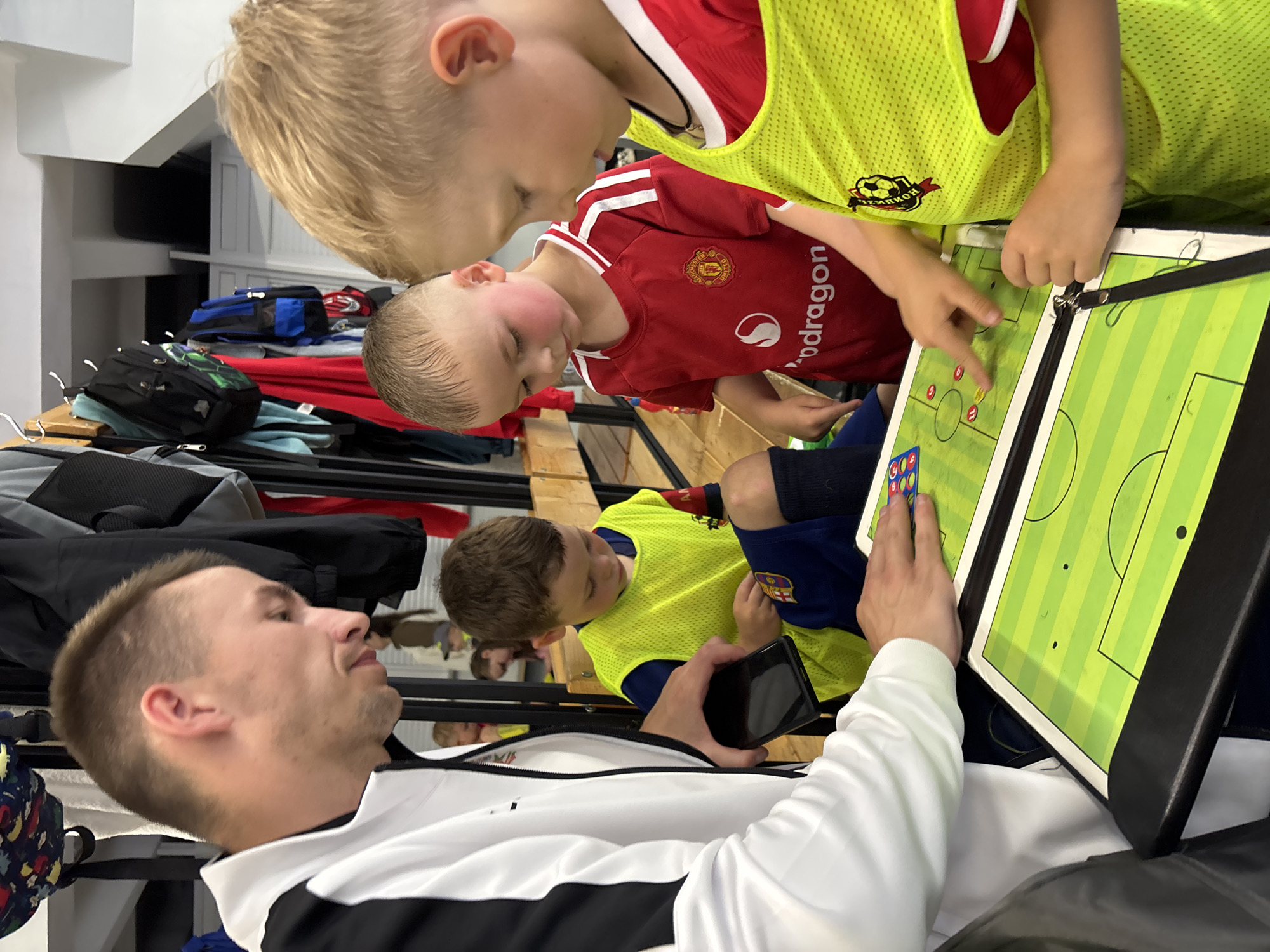 An adult male coach in a white and black jacket is standing to the left of the frame, looking down at a portable whiteboard depicting a soccer field. He holds a dark smartphone in his left hand and gestures with his right hand on the whiteboard. Two young boys, likely soccer players, are positioned in front of the whiteboard, also looking at it. The boy on the left wears a yellow soccer jersey over a blue shirt and shorts, while the boy on the right wears a red Manchester United soccer jersey. Both boys have blonde hair. The environment appears to be a locker room or indoor sports facility, with lockers, bags, and wooden benches visible in the background. The scene suggests a tactical discussion or coaching session. The specific location in Izhevsk, Russia, cannot be determined from the visual information provided in the image.