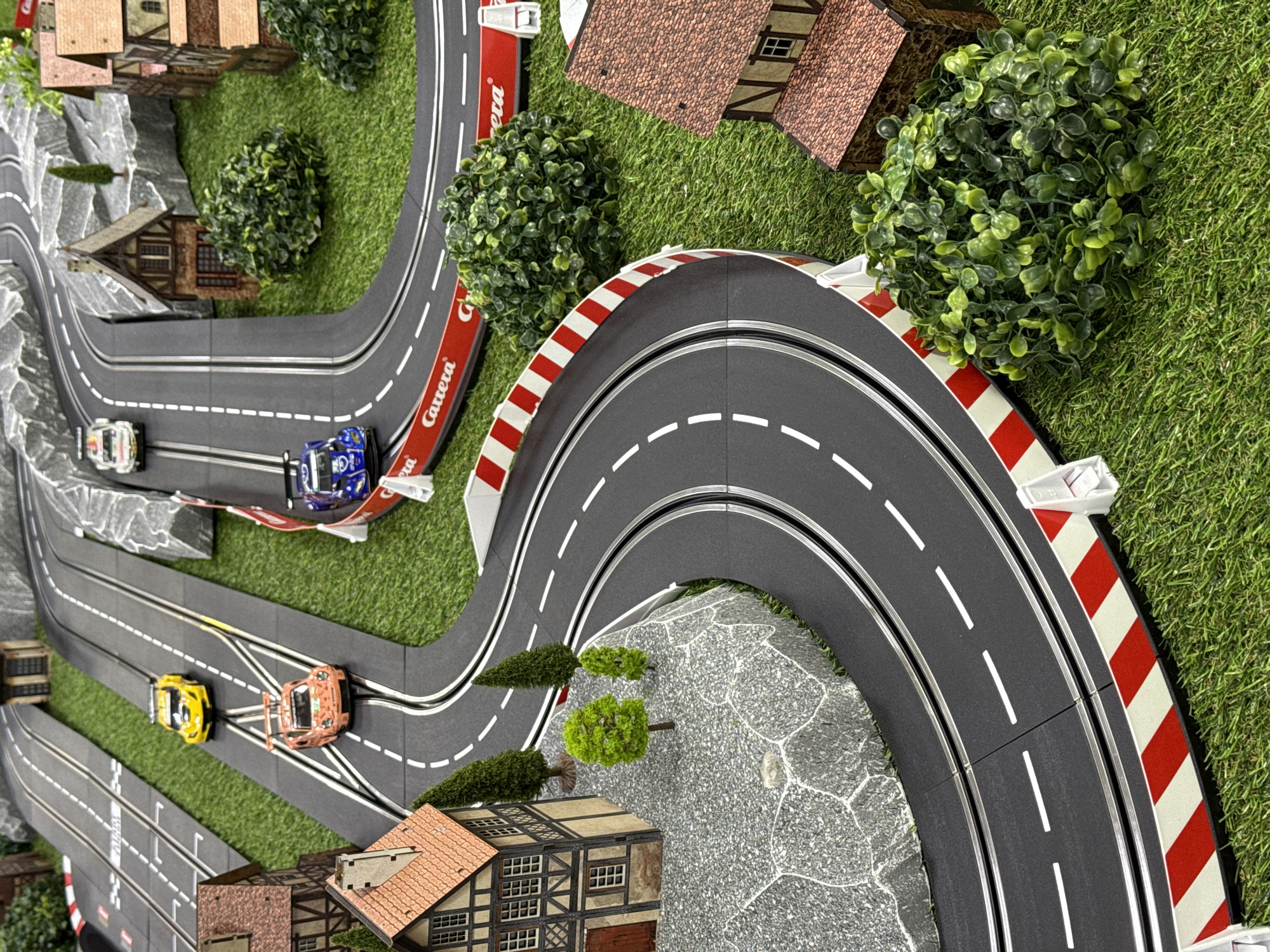 A miniature slot car racetrack is depicted with four cars currently on the track. The track winds through a landscape of faux green grass, miniature buildings, and a rock formation. The racetrack is marked with white lane lines and bordered by red and white barriers in some sections. Buildings are a European style with steeply pitched roofs and detailed facade. The miniature rock formation has a textured surface and is accented with tiny trees. The photograph is taken from a high-angle perspective, focusing on the layout of the racetrack and its surrounding scenery. The setting is a diorama, not a real location, and therefore, not in Sirius, Russia.