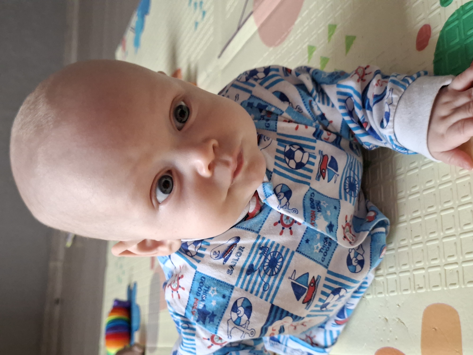 A close-up, medium shot depicts a baby, clad in a blue and white "sailor"-themed pajama set, lying on a patterned playmat. The baby's head is bald, and the gaze is directed slightly upwards towards the camera. The playmat is made of interlocking foam tiles. A colorful toy, possibly a stacking rainbow toy, is partially visible in the lower left corner of the frame. The location and city are unknown.