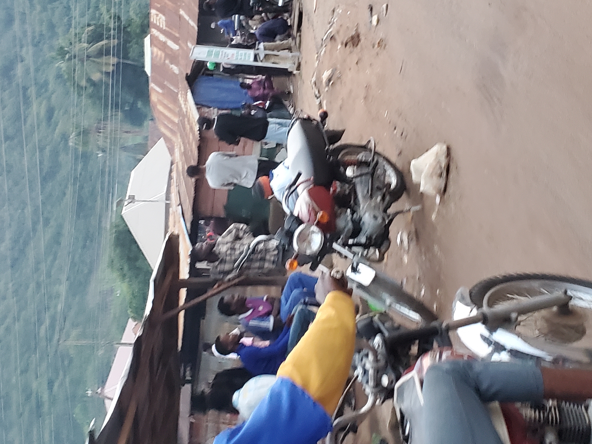 The image depicts an outdoor street scene in Mararaba, Nigeria, characterized by a reddish-brown earth ground. Approximately ten adult males are visible across the midground. In the immediate foreground on the left, a person's torso and arm, clothed in a blue and yellow top, is seen holding the handlebars of a partially visible red and white motorcycle.
In the midground, several individuals are gathered. Two men are seated to the left, one wearing a blue top and the other a purple top. A red and black motorcycle is parked facing left in the center-midground. To the right, various men are standing, one in a plaid shirt, others in casual attire, near makeshift structures with corrugated iron roofs, some visibly rusted. A white vertical signpost stands upright on the right side. On the dusty ground, a crumpled white plastic bag is located near the parked motorcycle.
The background features additional buildings with varied roofs, including both modern white and rusted corrugated iron. Behind these structures, a dense green vegetated hillside forms the horizon. Multiple thin power lines are horizontally stretched across the upper midground. Several palm trees are discernible against the green background, and a faint haze or smoke is present in the distant air.